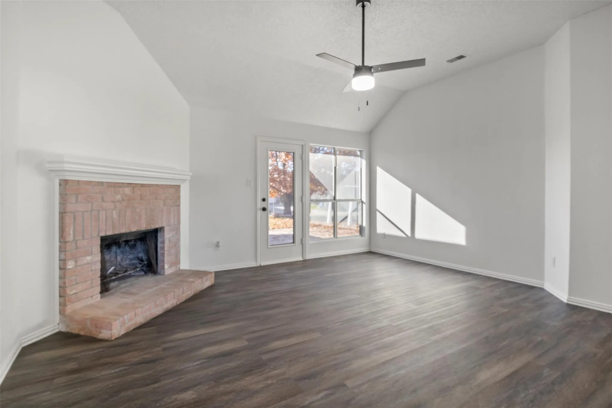 Vaulted ceiling with a ceiling fan, brick fireplace with a hearth, wood-finish flooring, and a glass-paned exterior door