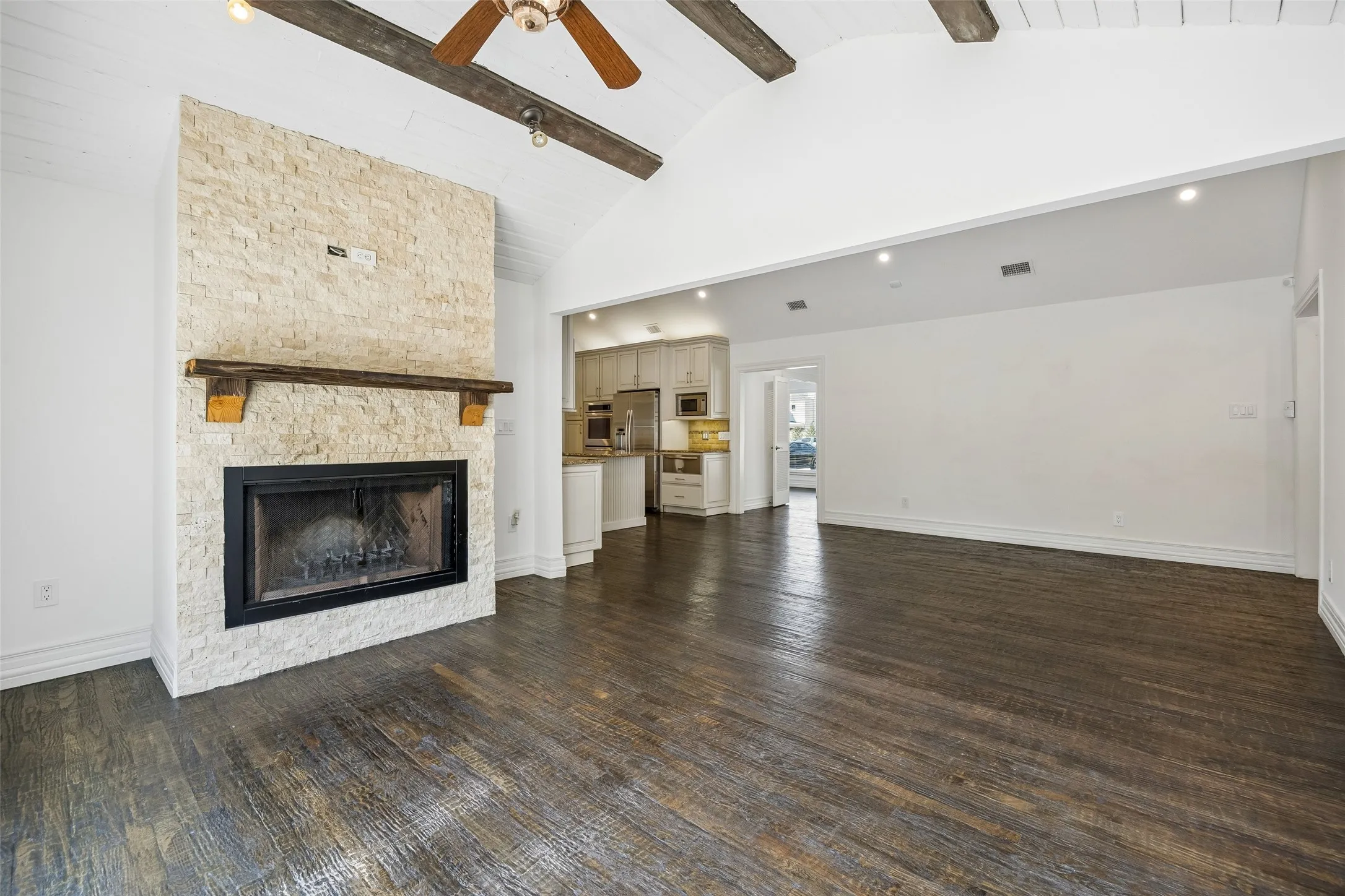 Vaulted ceiling with exposed wood beams, wood-finish flooring, stone-clad fireplace with wood mantel, recessed lighting, and a ceiling fan
