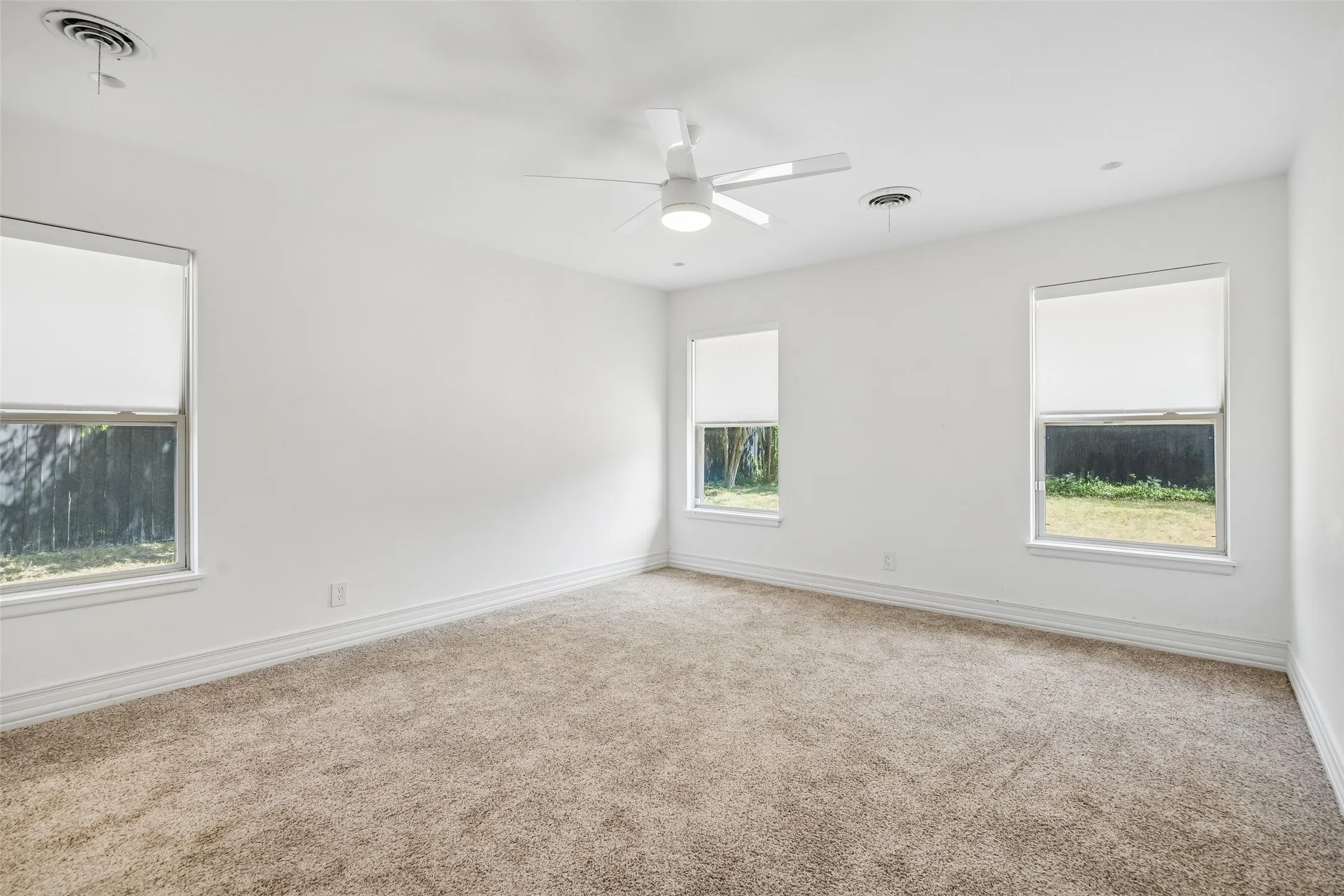 Neutral-toned carpet flooring, crisp white walls, three windows with roller shades, and a ceiling fan with integrated lighting