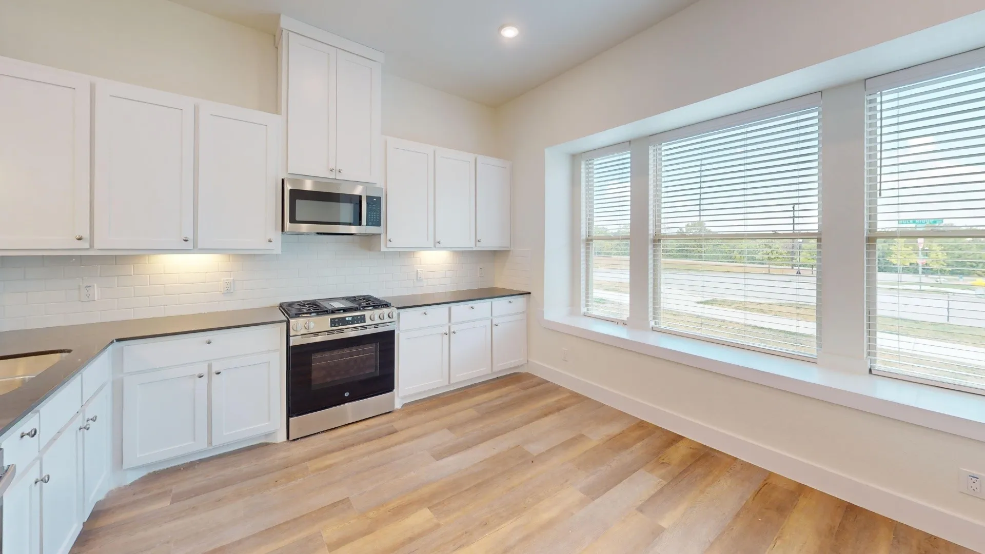 White shaker cabinetry, stainless steel appliances, subway tile backsplash, dark countertops, and wood-finish flooring