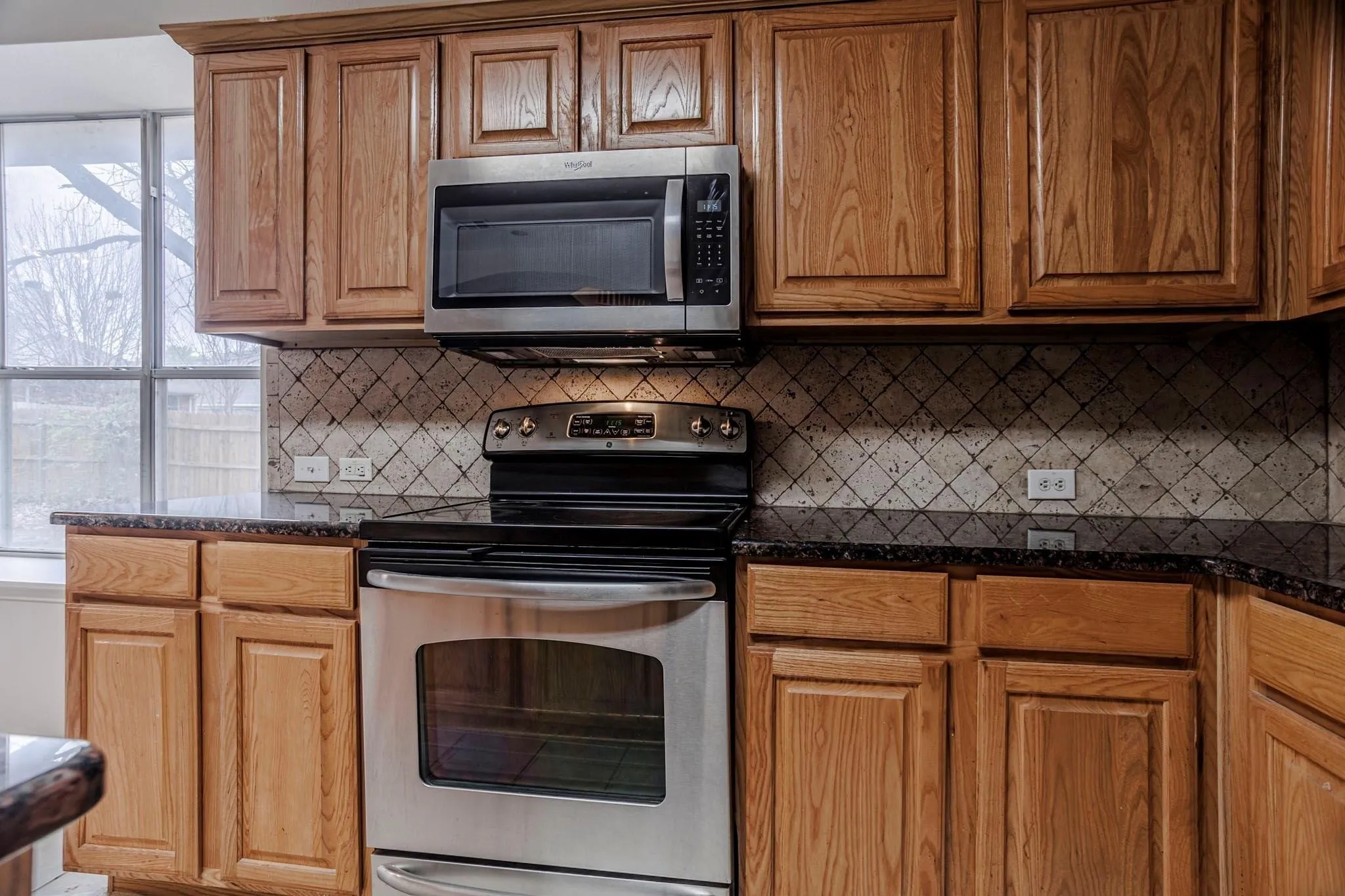 Wood-finish cabinetry, stainless steel range, stainless steel microwave, dark speckled countertops, and a decorative tile backsplash