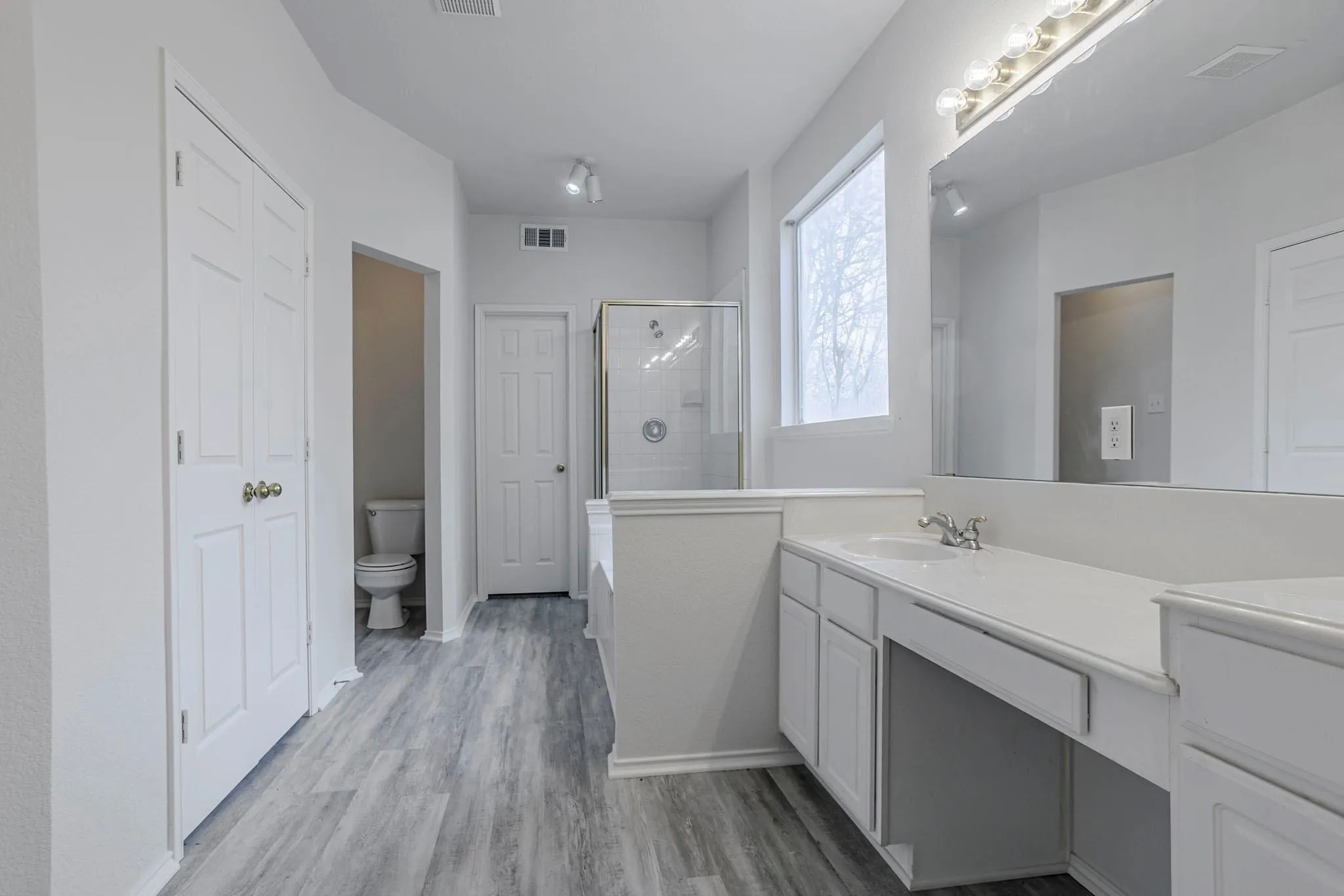 Wood-finish flooring, white vanity with integrated sink, large vanity mirror, glass-enclosed shower, and a private toilet area