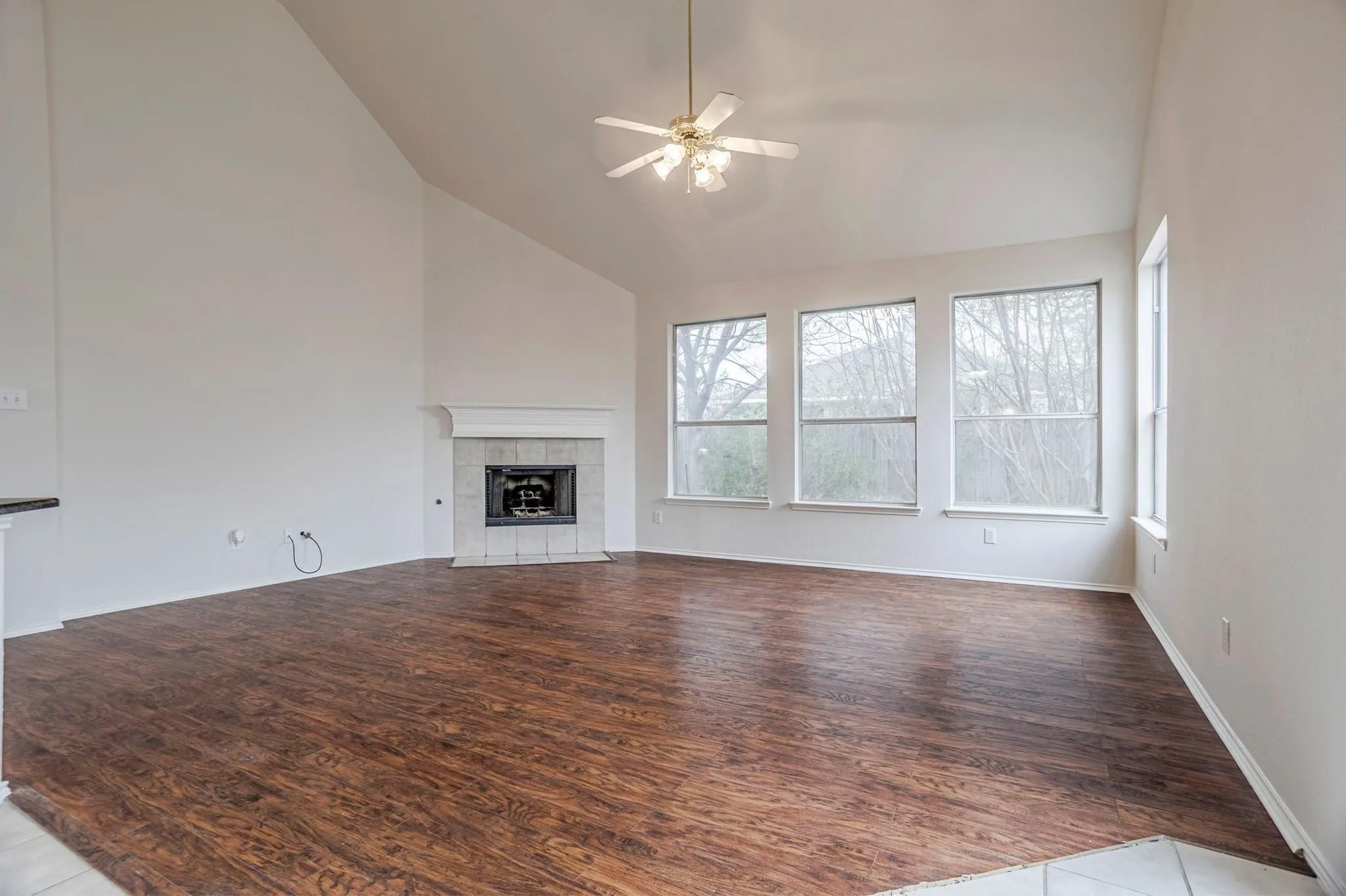 Vaulted ceiling with a ceiling fan, wood-finish flooring, tiled fireplace with mantel, and three windows