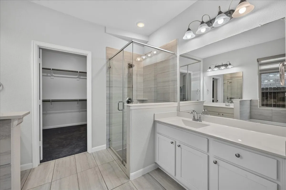 Dual vanity with white cabinetry, a glass-enclosed shower, a large wall mirror, and light-toned tile flooring