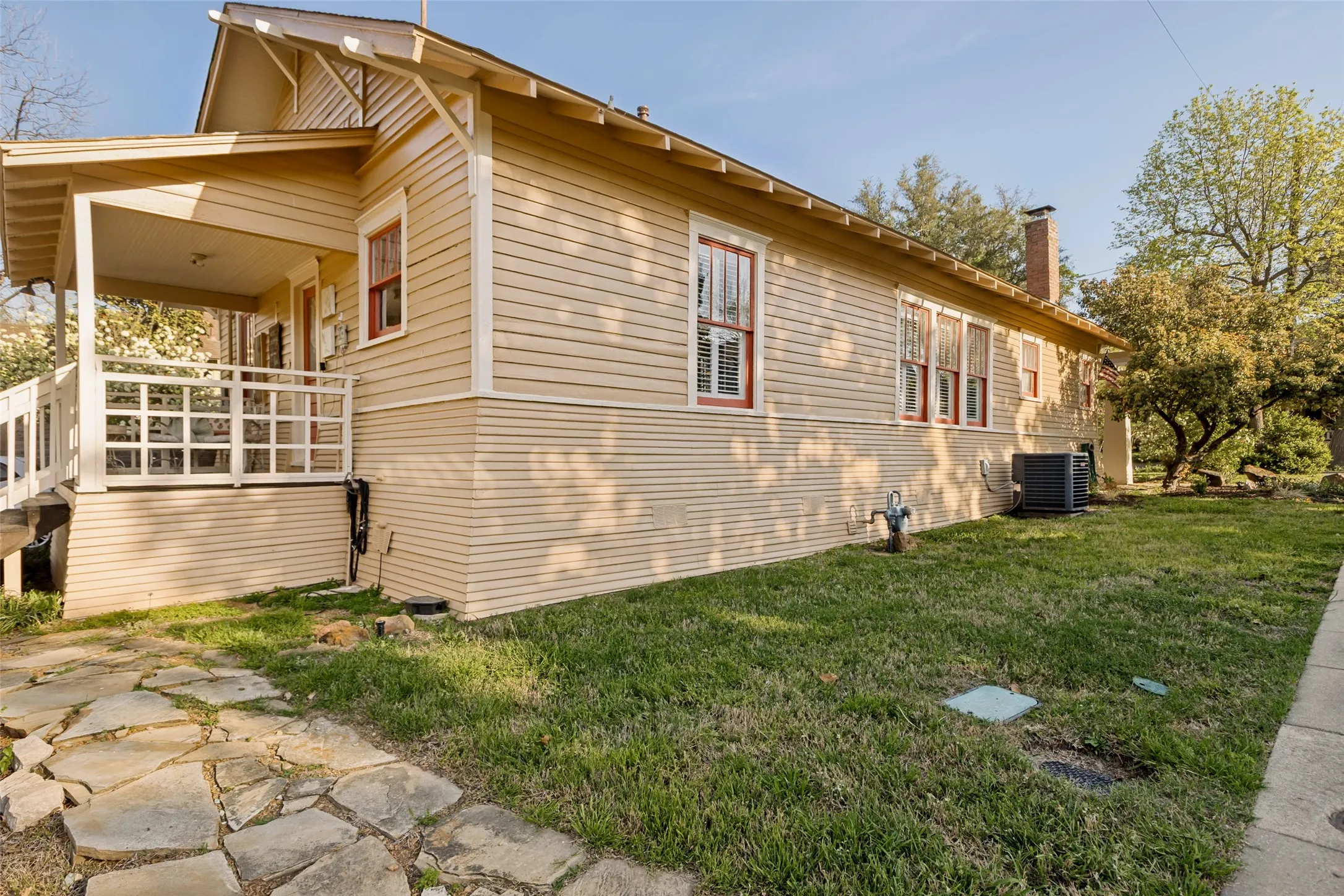 Exterior featuring a covered porch with white railings, tan horizontal siding, multiple windows with red trim, a visible chimney, and a flagstone pathway