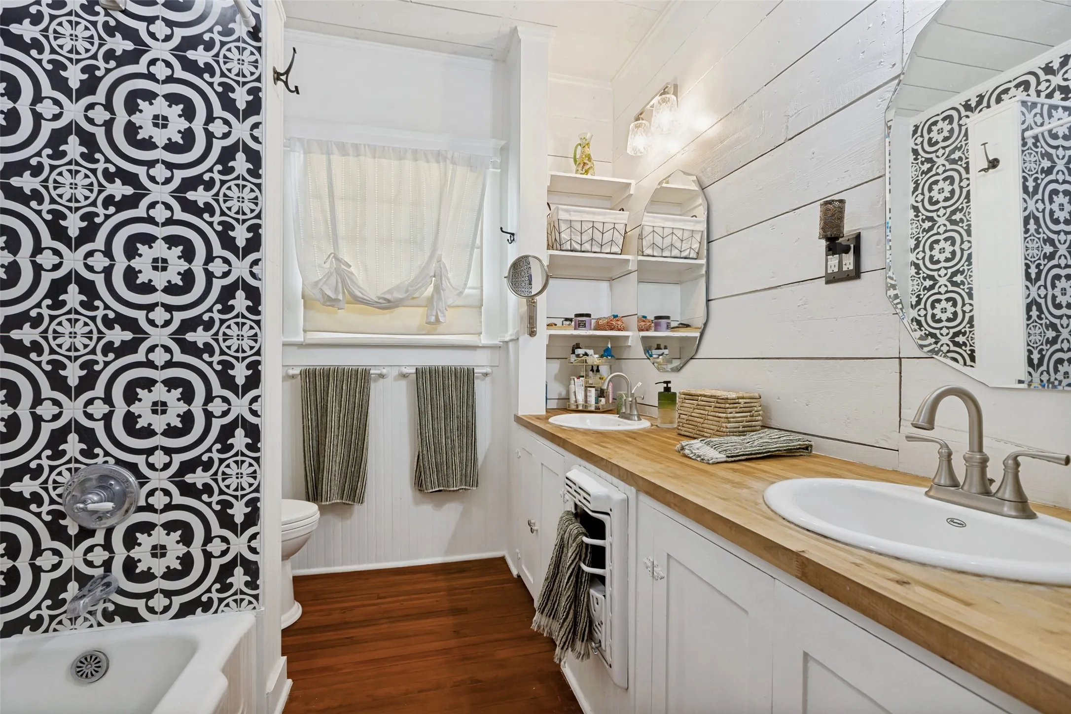 Dual vanity with wood-finish countertop, decorative tile shower surround, wood-finish flooring, and shiplap wall paneling