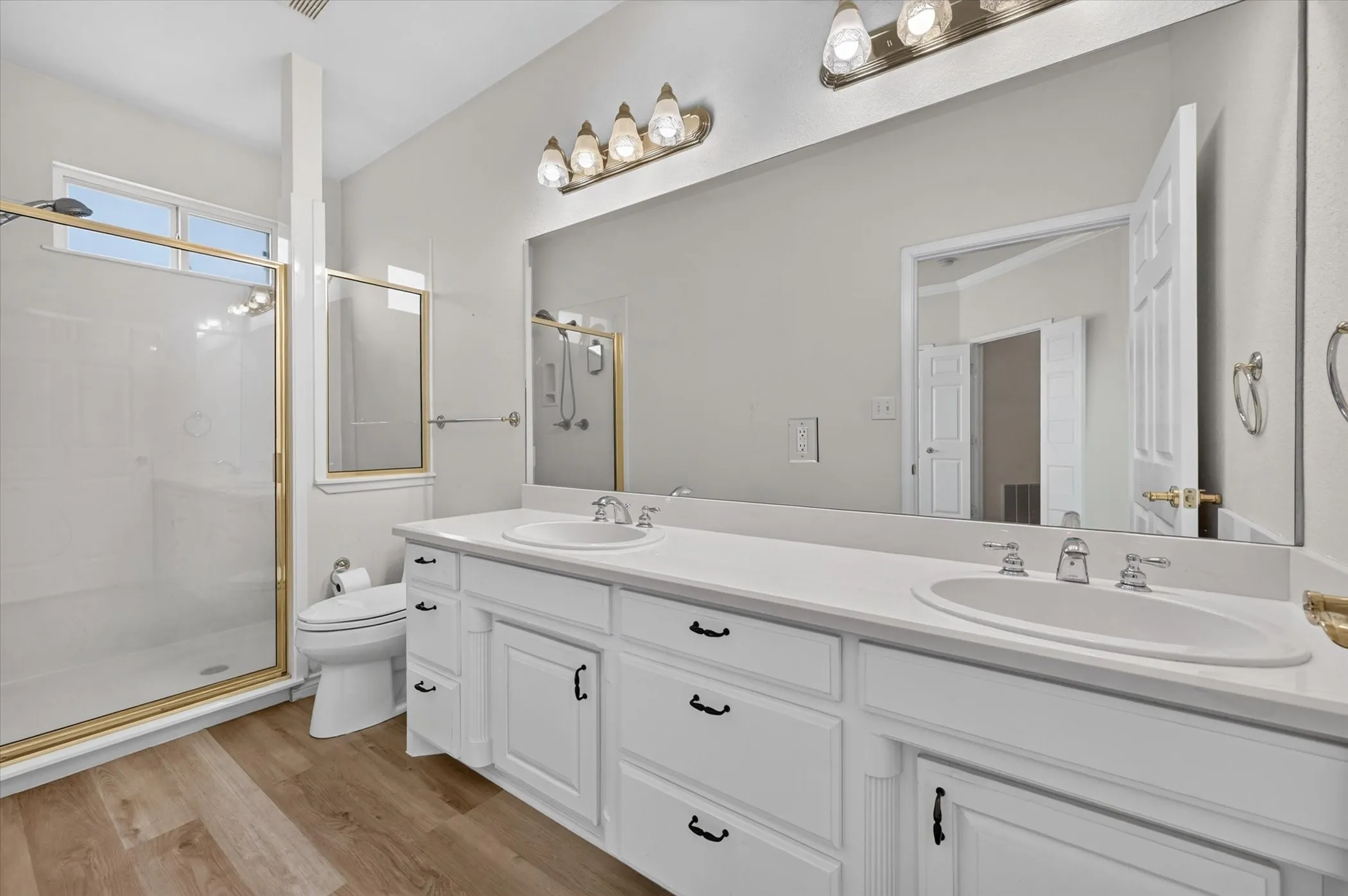 Spa-inspired primary bathroom featuring dual vanities with white cabinetry and brushed nickel hardware, expansive mirror with integrated lighting, quartz countertops, and a walk-in shower with frameless glass enclosure and tile surround