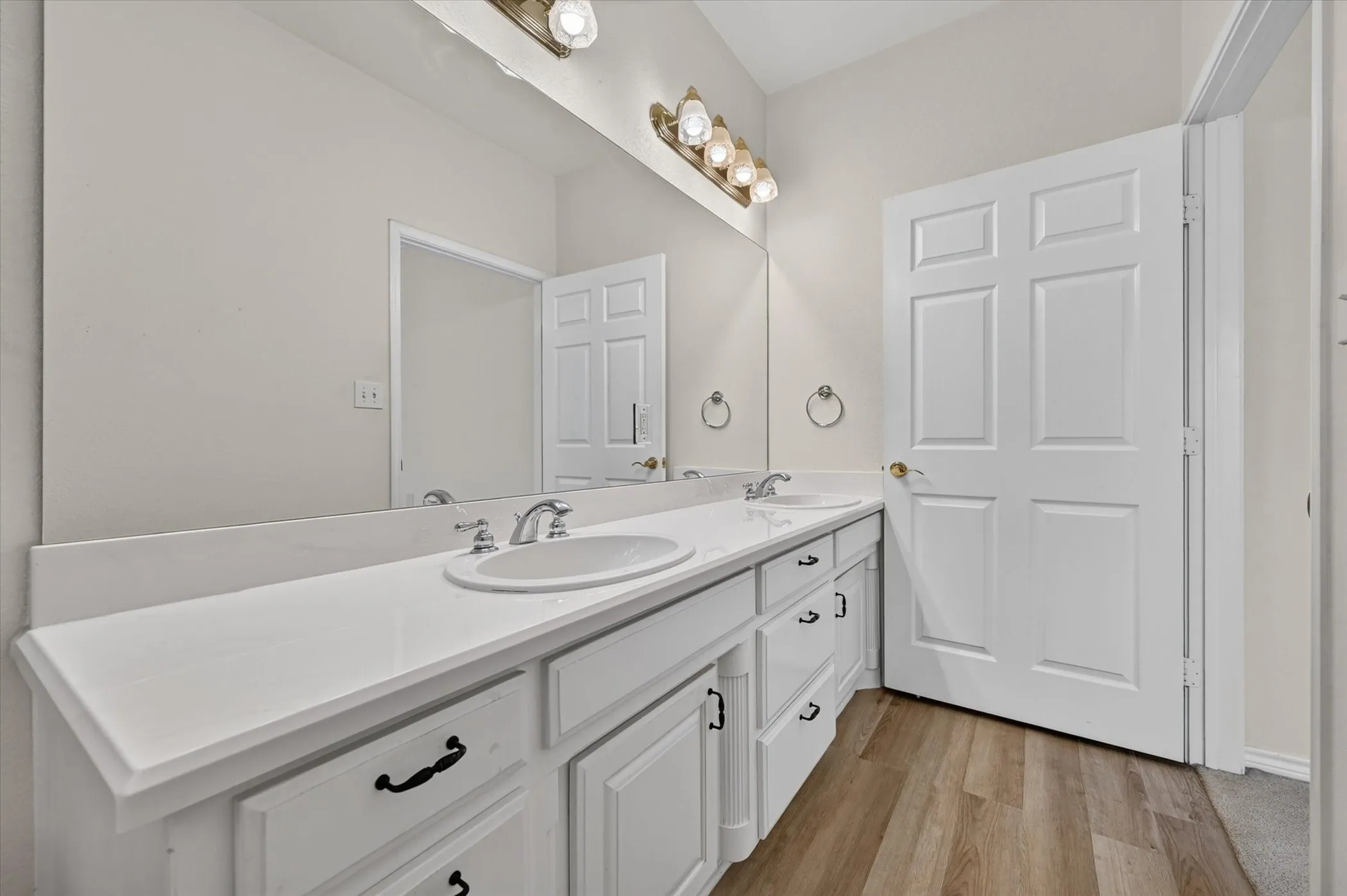 Double vanity with white cabinetry and integrated sinks, brushed nickel fixtures, wood-finish flooring, and a large wall-mounted mirror