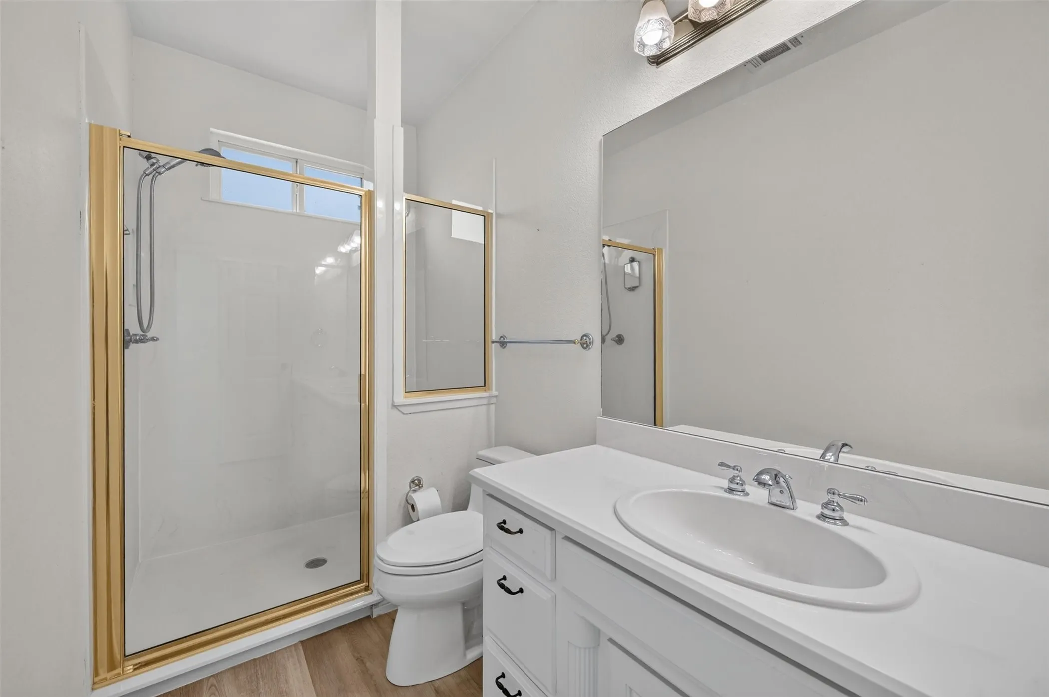 Bathroom featuring a vanity with an integrated sink, a large mirror, a shower with a gold-finish frame, and wood-finish flooring