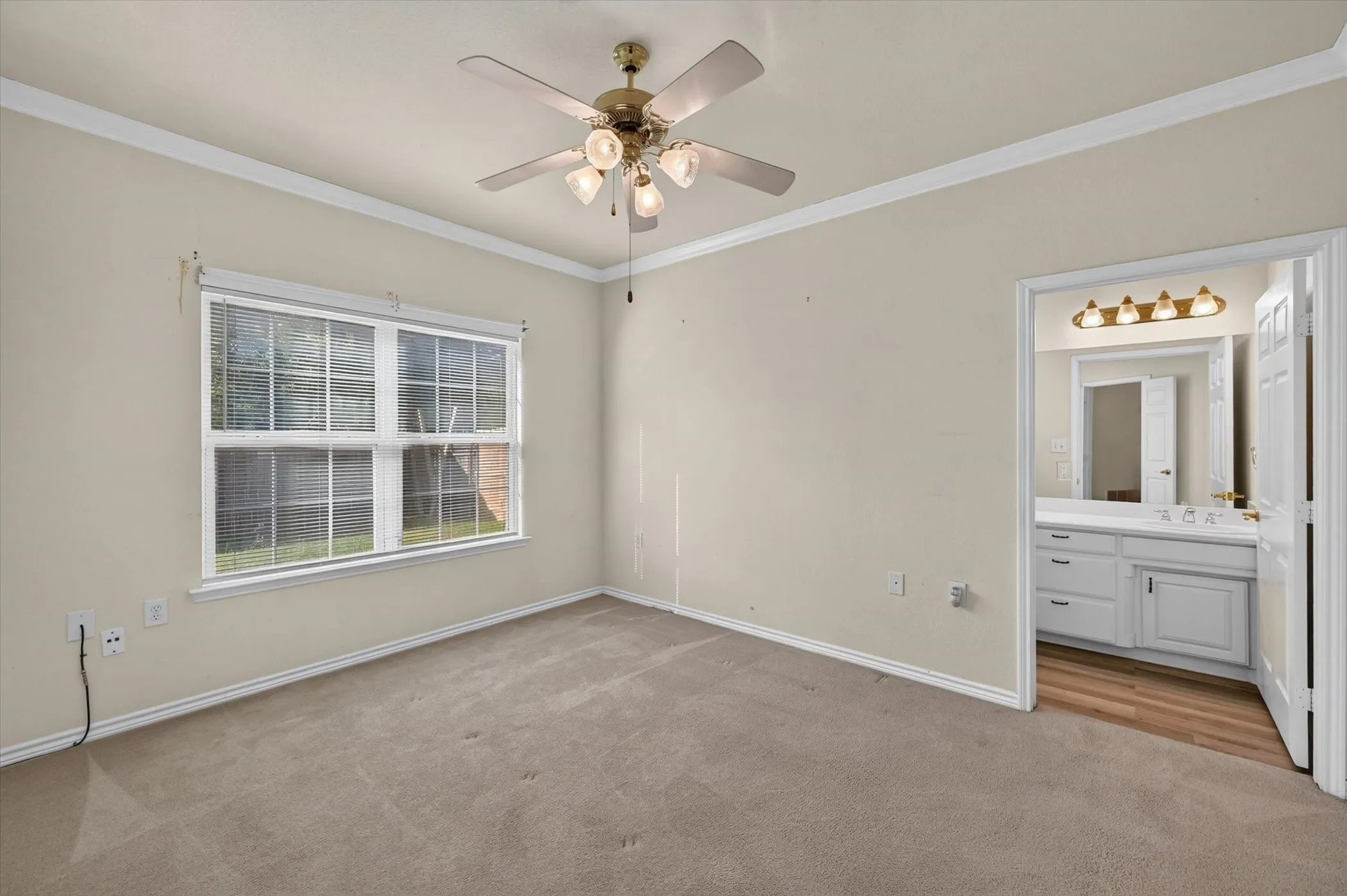 Spacious primary bedroom featuring a large double-hung window with multiple panes, ceiling fan with integrated lighting, hard-wood flooring, and an ensuite bathroom with dual-sink vanity, framed mirror with integrated lighting, and white cabinetry