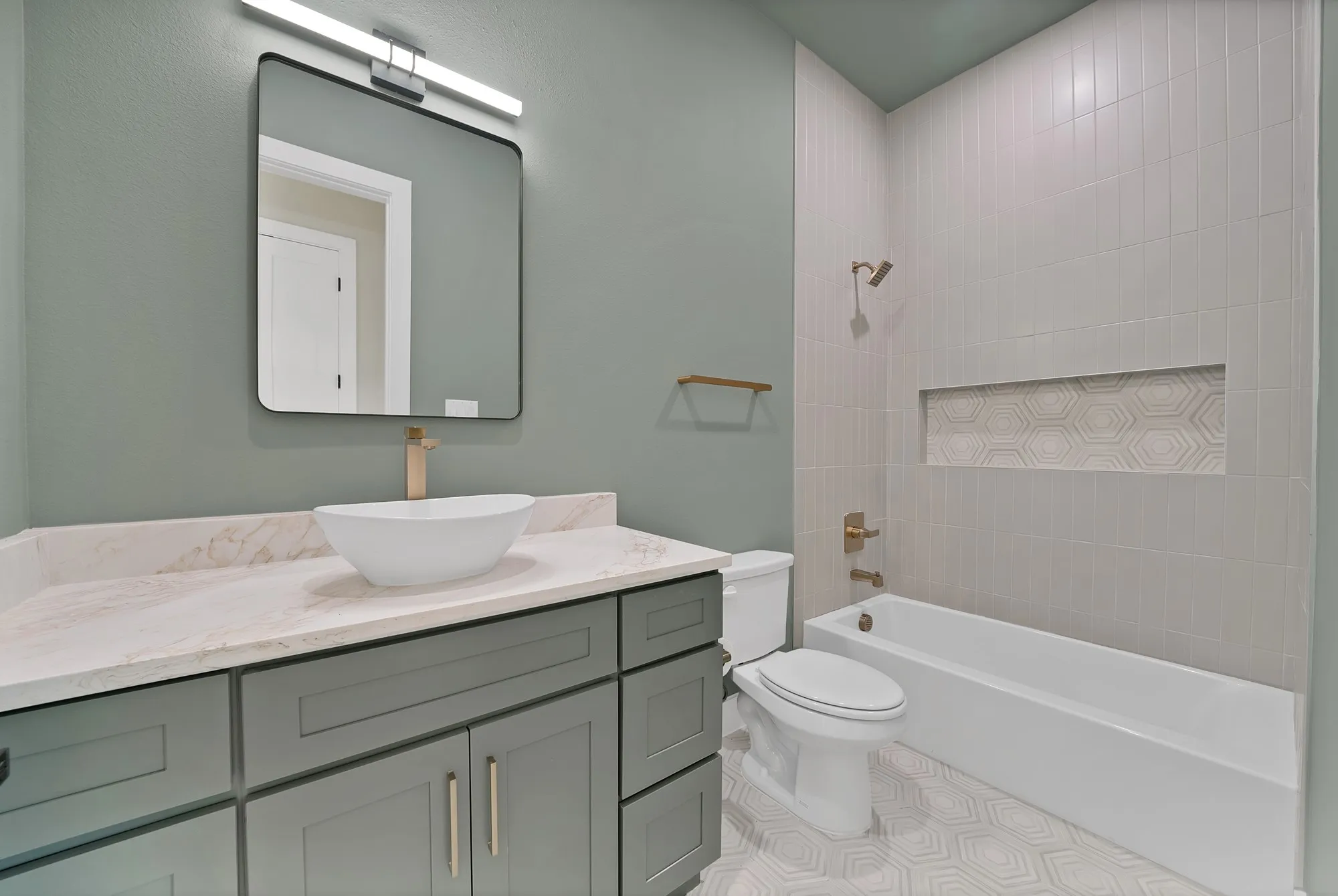 Vanity with a vessel sink and gold-tone faucet, framed mirror with integrated lighting, built-in bathtub with shower, tiled shower surround with a decorative niche, and patterned floor tile