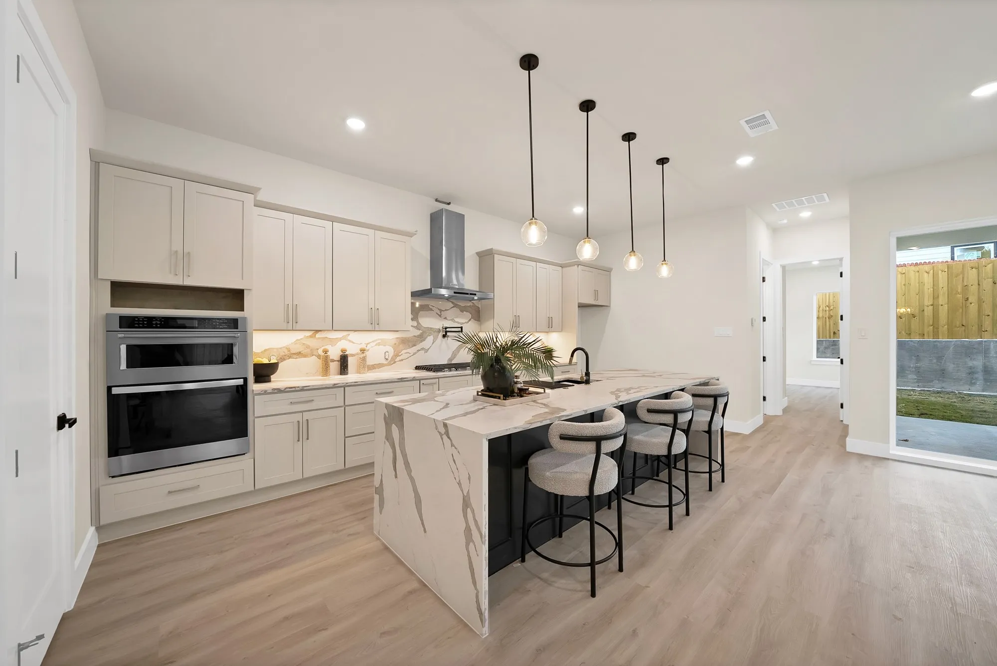 Gourmet kitchen featuring a large waterfall island, stainless steel double ovens, a built-in range hood, light wood-finish flooring, and pendant lighting