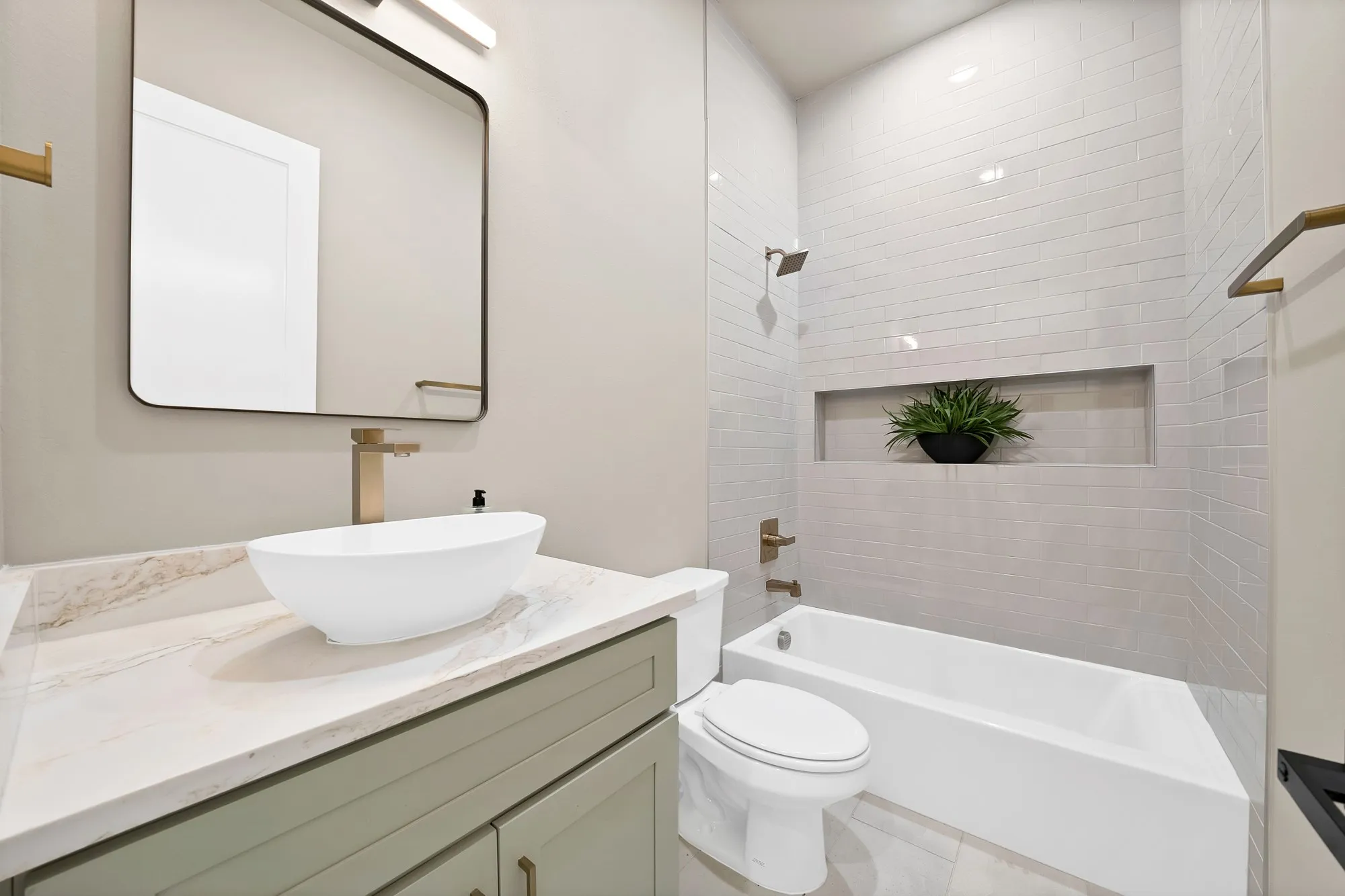 White vessel sink with brass-finish faucet, framed mirror with integrated lighting, green-finish vanity with brass-finish hardware, subway-tiled shower with recessed niche, and a white bathtub