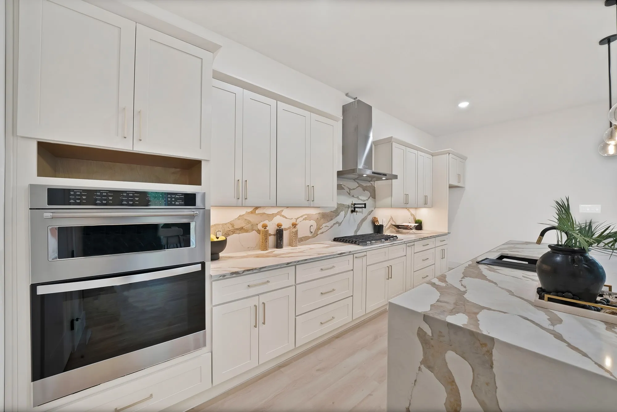 Built-in stainless steel double oven, white shaker-style cabinetry, patterned countertop and backsplash, stainless steel range hood, and light wood-finish flooring
