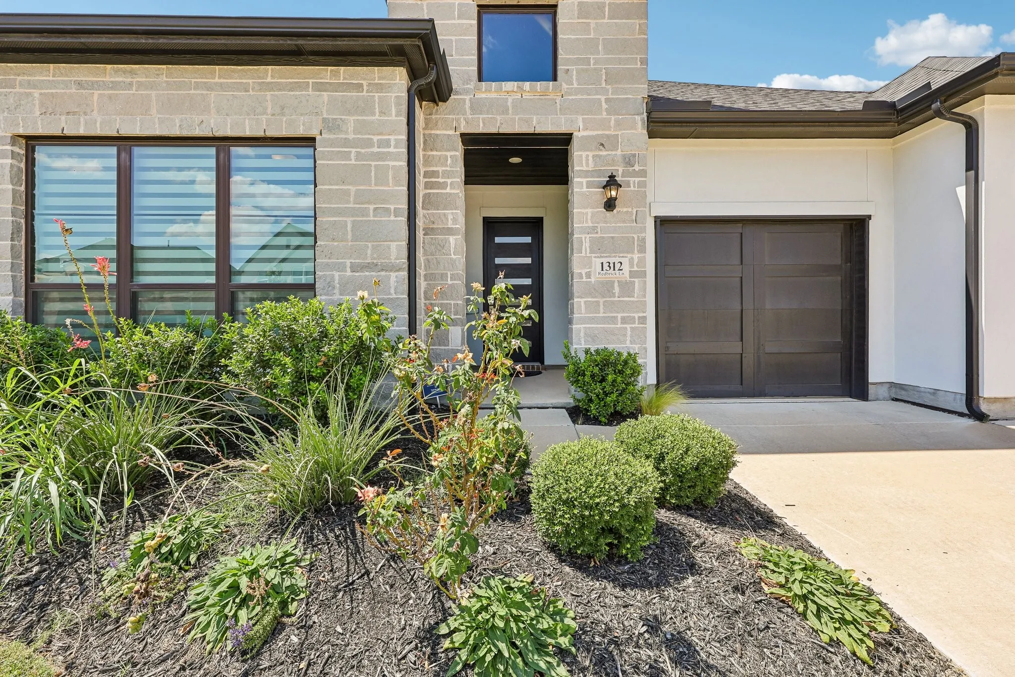 Stone façade with dark trim accents, modern entry door, integrated garage, and mature landscaping