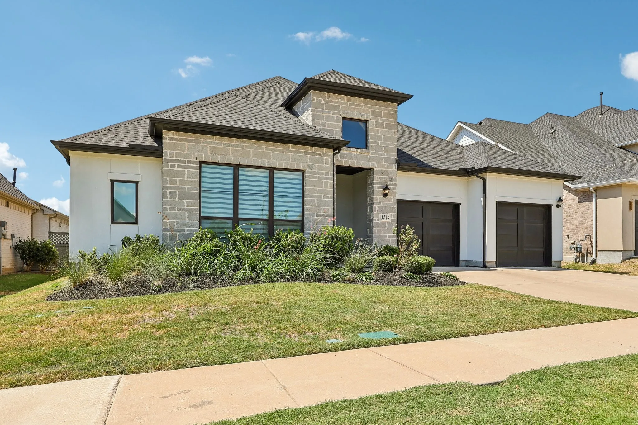 Modern exterior featuring stone and stucco finishes, a dark shingle roof, two dark-toned garage doors, and a concrete driveway