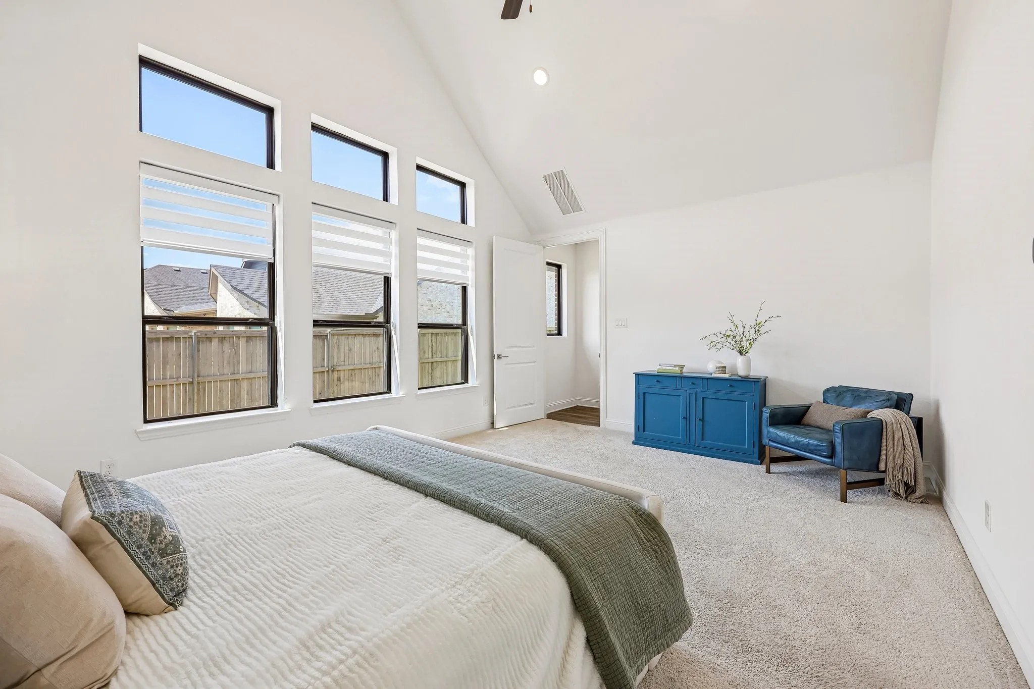 Vaulted ceiling, multiple windows with blinds, light-colored carpeting, white walls, and a ceiling fan