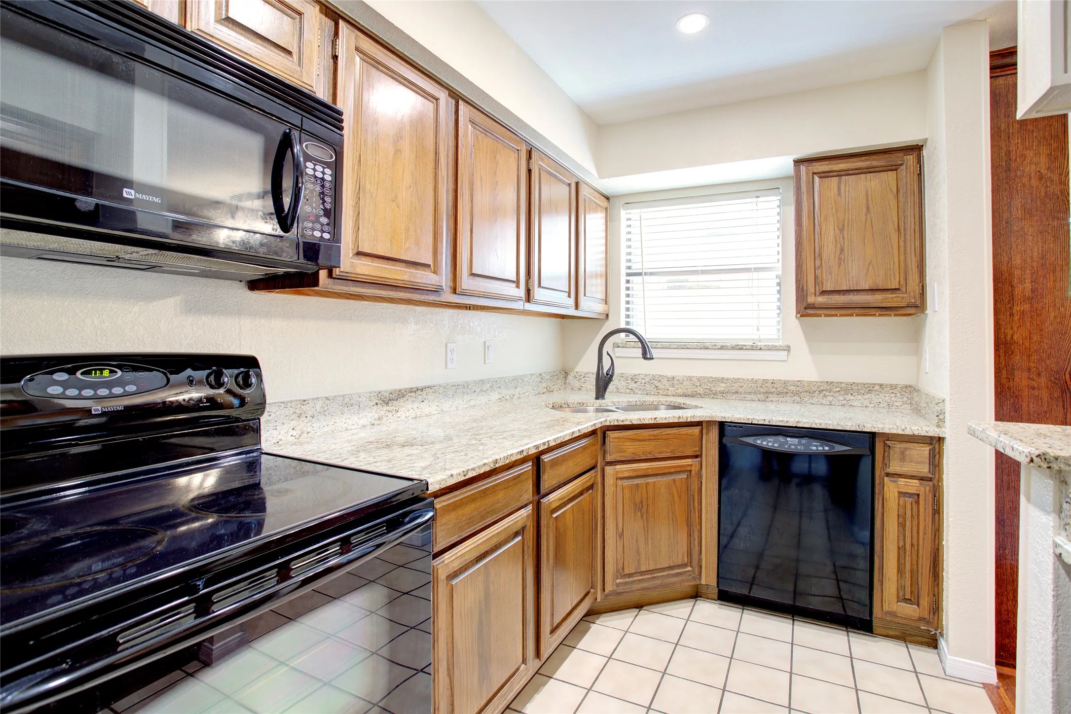 Wood cabinetry, light-toned granite-finish countertops, black appliances, and tile flooring