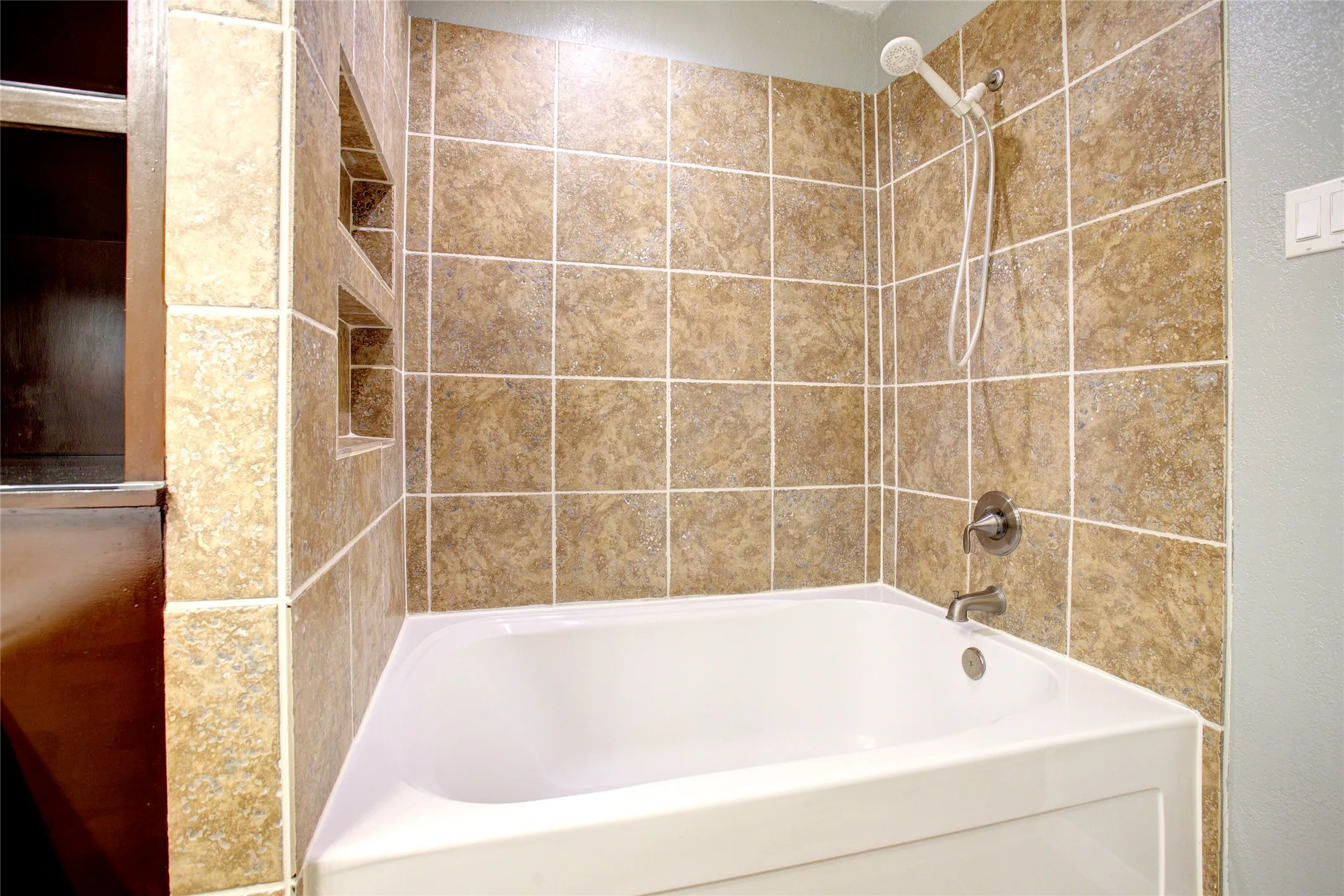 Built-in alcove shelving, ceramic tile surround, white tub, brushed nickel fixtures, and a handheld showerhead