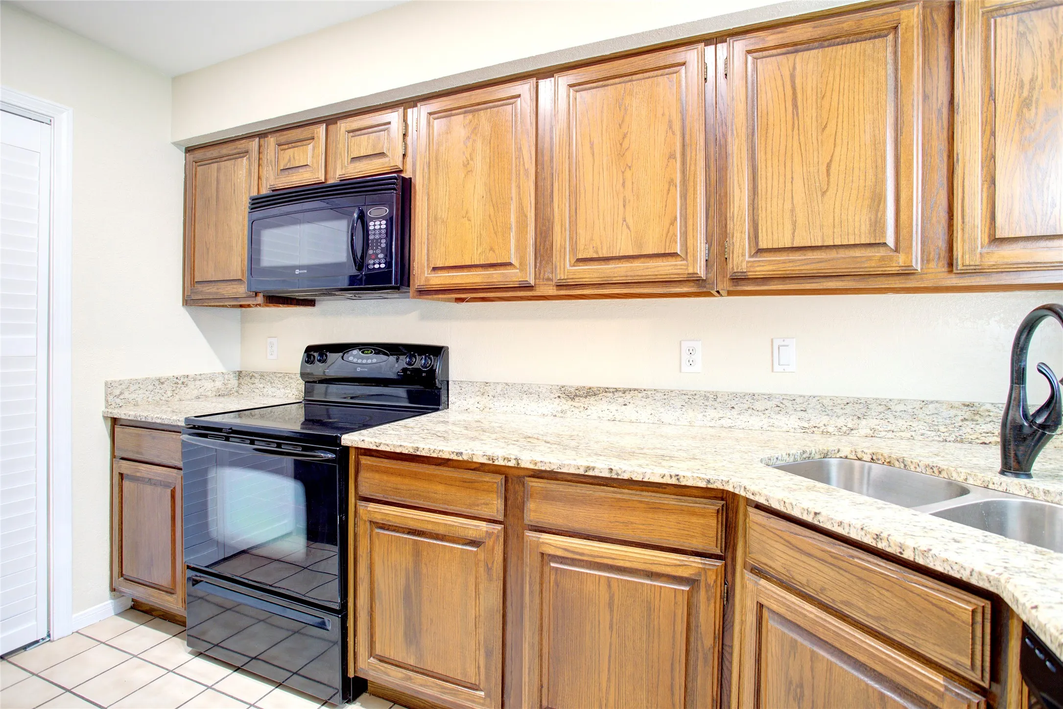 Wood-finish cabinetry, light-toned countertops, tile flooring, black range, and dual stainless-steel sink