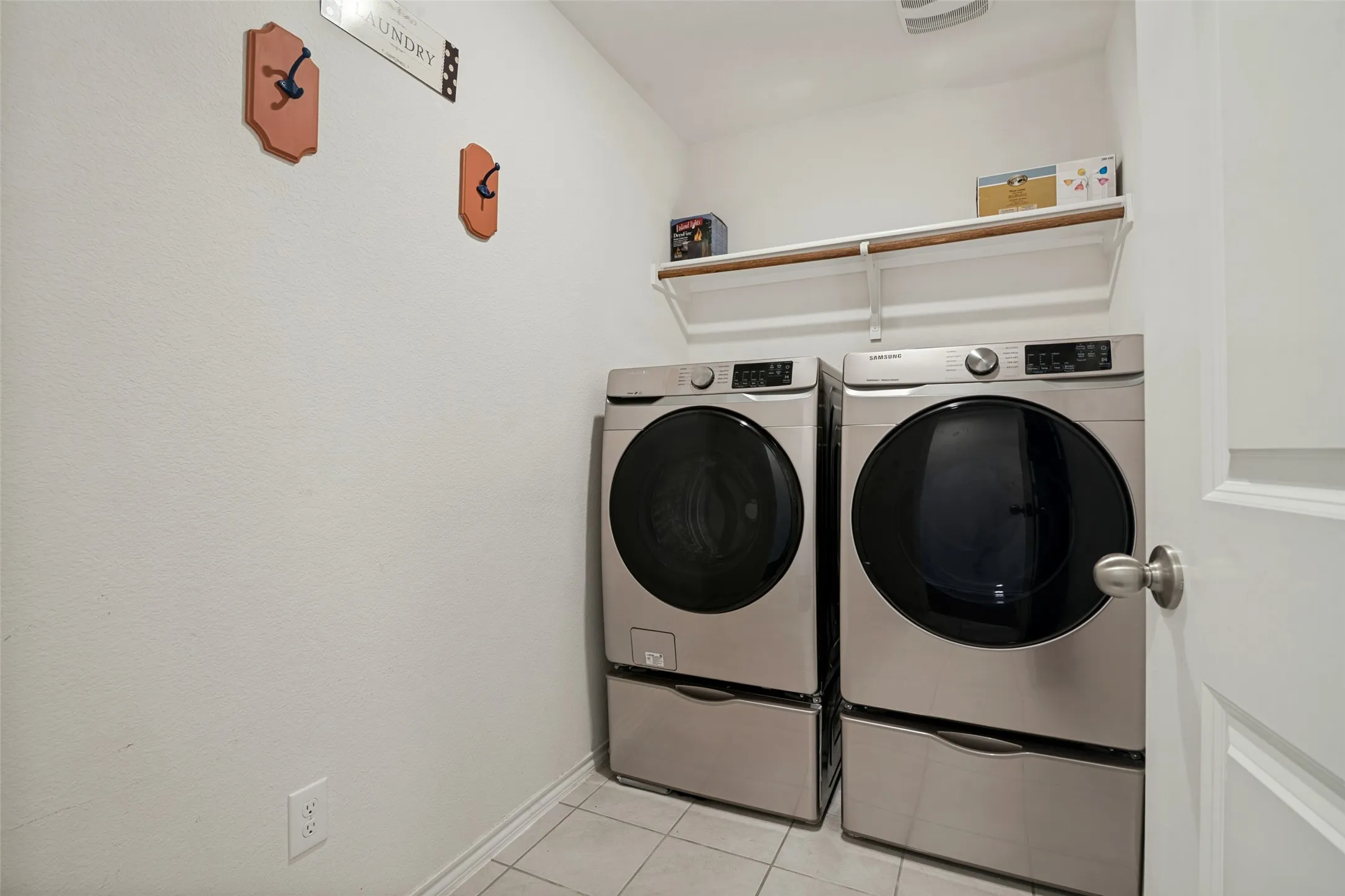 Laundry area with a front-loading washer and dryer, elevated on pedestals, complete with an overhead storage shelf and ceramic tile flooring
