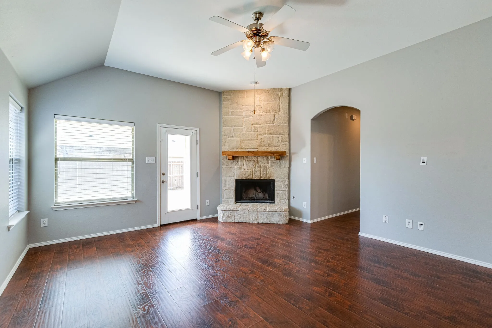 Vaulted ceiling, wood-finish flooring, stone-clad fireplace with wood mantel, ceiling fan with integrated lighting, and an arched doorway