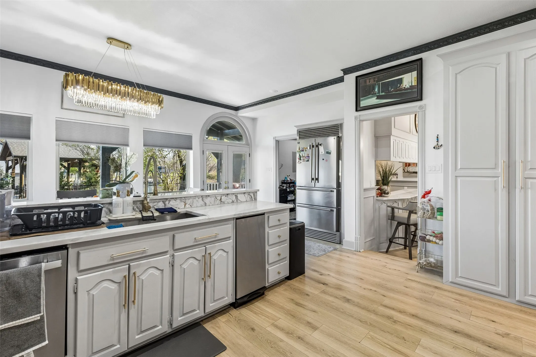 Kitchen featuring light wood-finish flooring, white cabinetry, integrated stainless steel appliances, a center island with sink, and a modern linear chandelier
