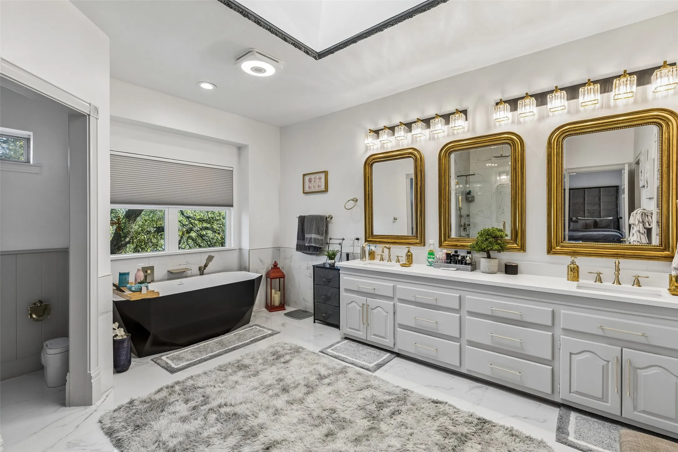 Freestanding soaking tub, dual vanity with two sinks, gold-framed mirrors, vanity lighting, and white tile flooring