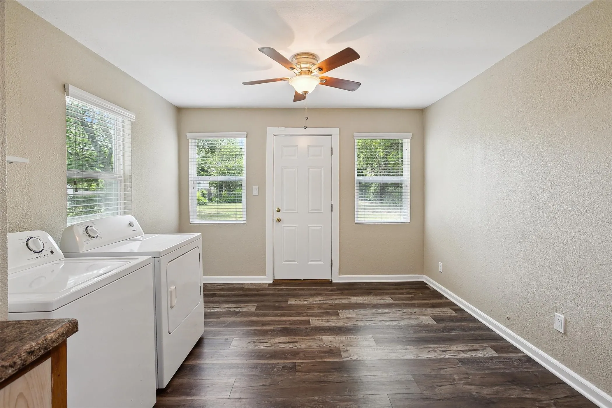 Laundry area with laminate flooring, a ceiling fan, door leading to backyard and white window blinds