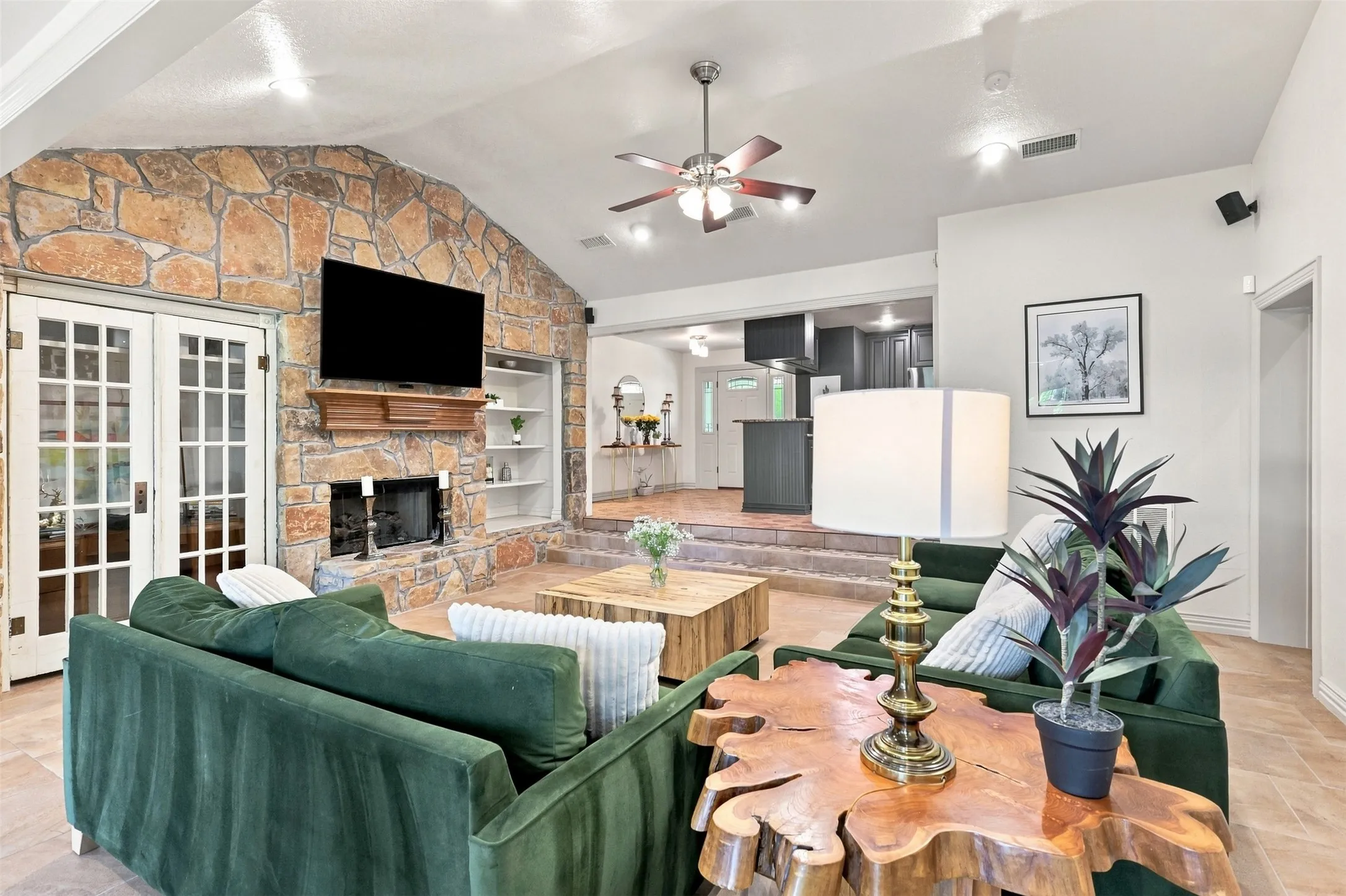 Vaulted ceiling featuring a ceiling fan, stone fireplace with a wood mantle, built-in shelving, and double French doors with glass panels