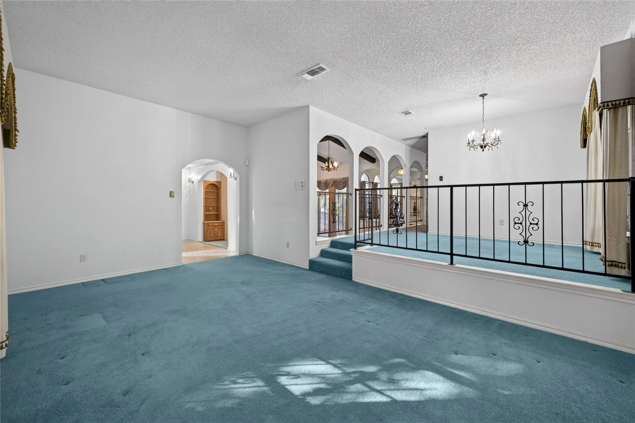 Expansive room featuring blue carpet, a decorative iron railing, an arched entryway, and a chandelier