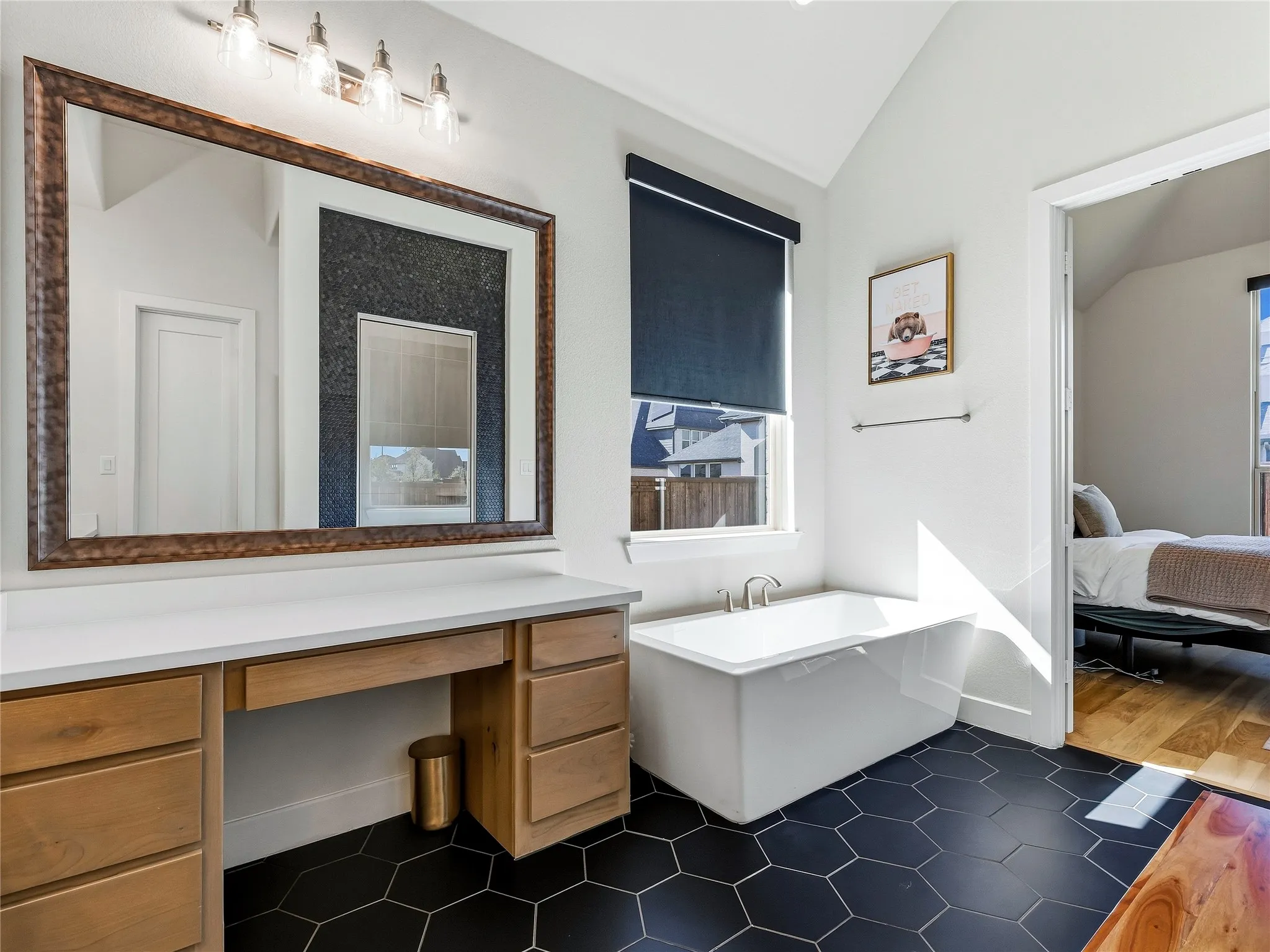 Freestanding soaking tub, hexagon floor tiling, wood-finish vanity with countertop, large framed mirror, and vaulted ceiling
