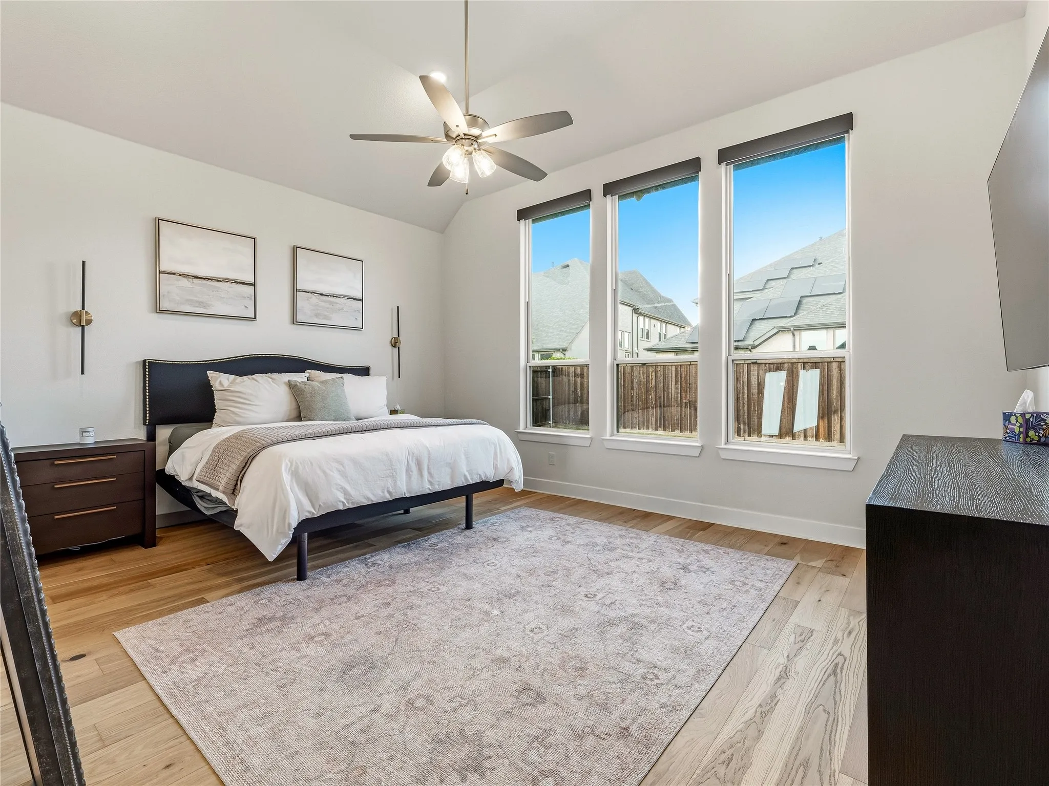 Vaulted ceiling with a ceiling fan, three tall windows, wood-finish flooring, and wall-mounted sconces