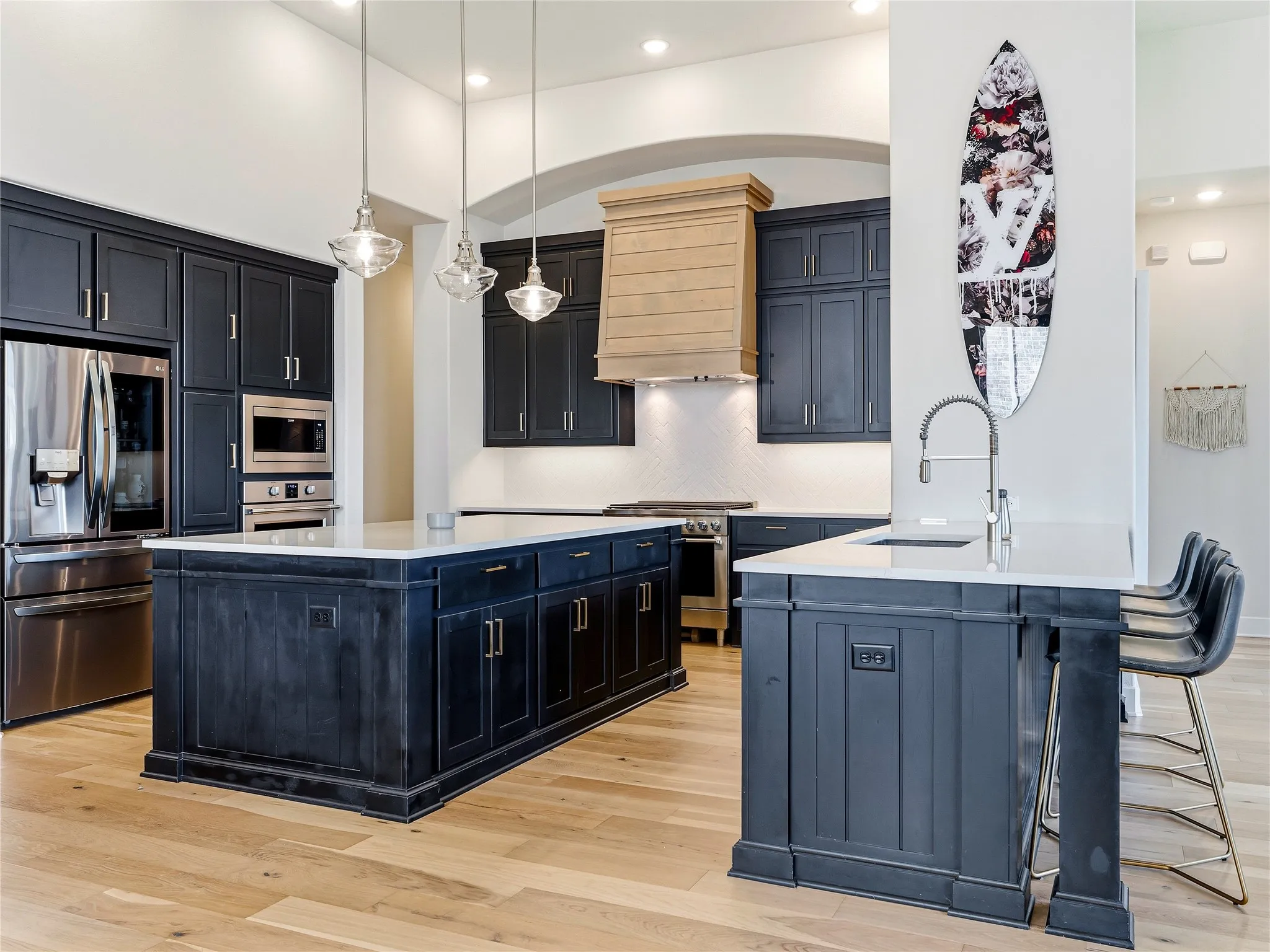 Kitchen featuring dark-toned cabinetry, light-toned wood-finish flooring, stainless steel appliances, dual islands with white countertops, and a wood-finish range hood