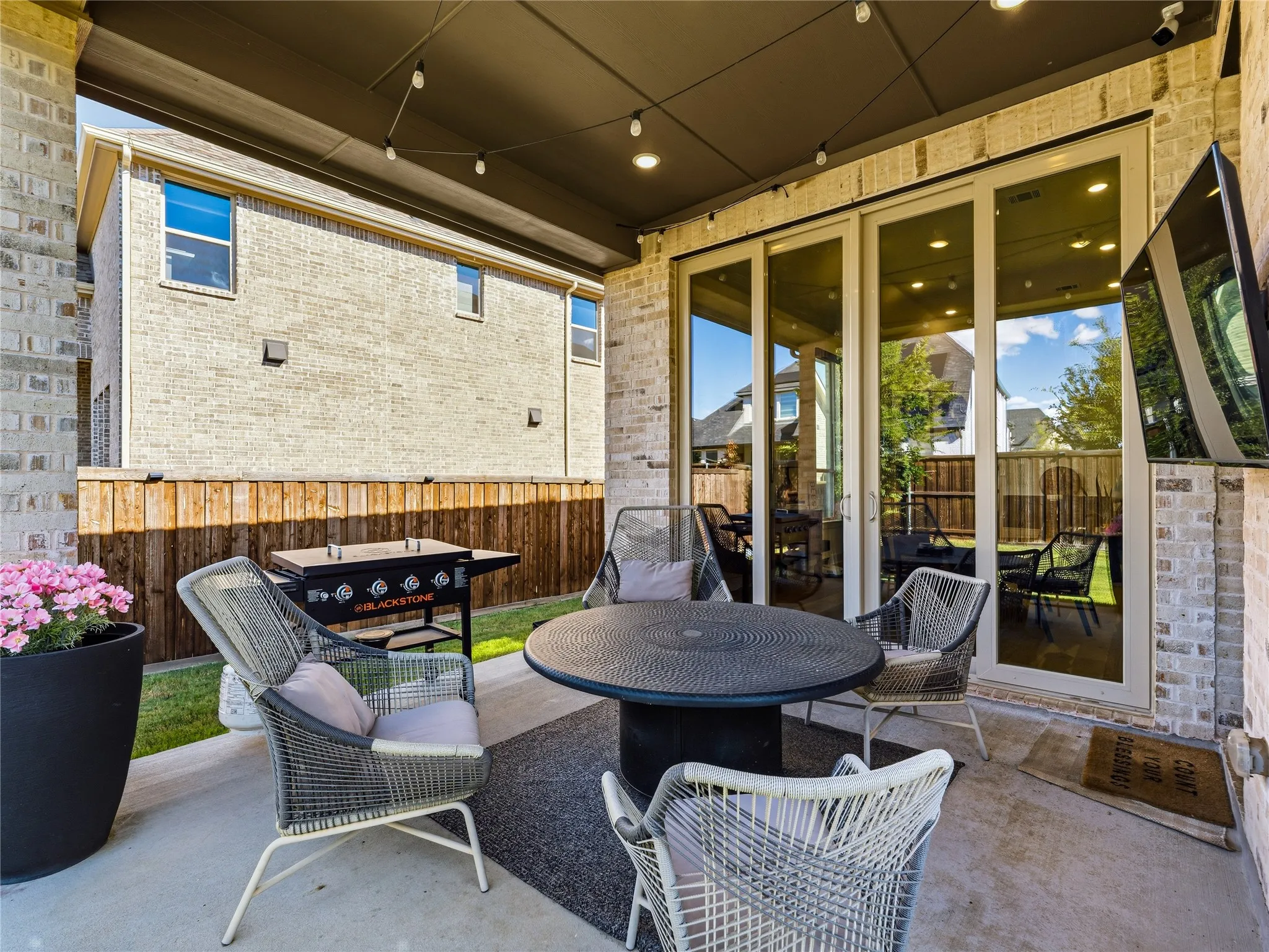 Covered patio with a mounted television, string lighting, brick columns, and a full wall of glass doors