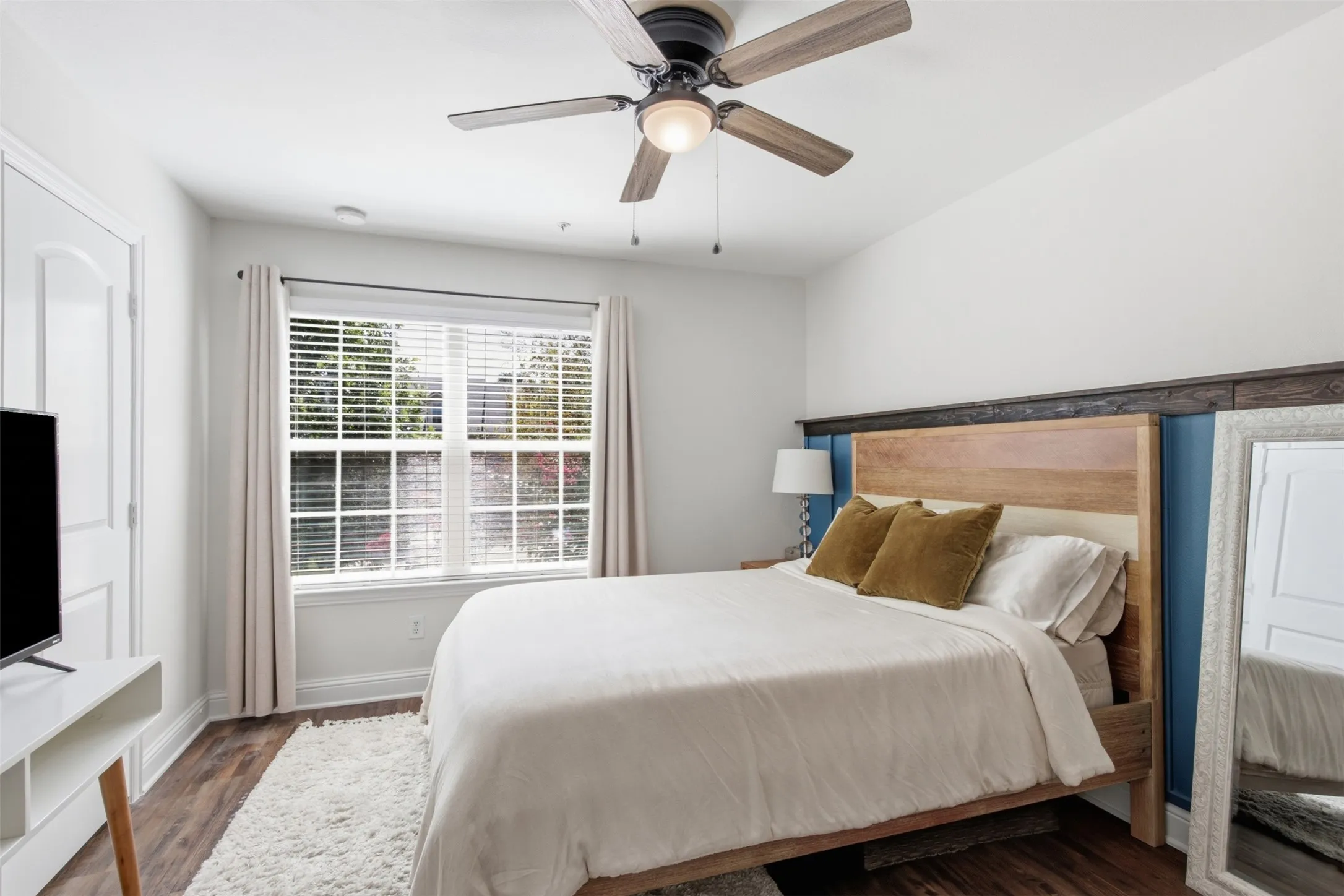 Hardwood-finish flooring, ceiling fan with integrated lighting, window with blinds and drapes, white interior door, and built-in wall paneling