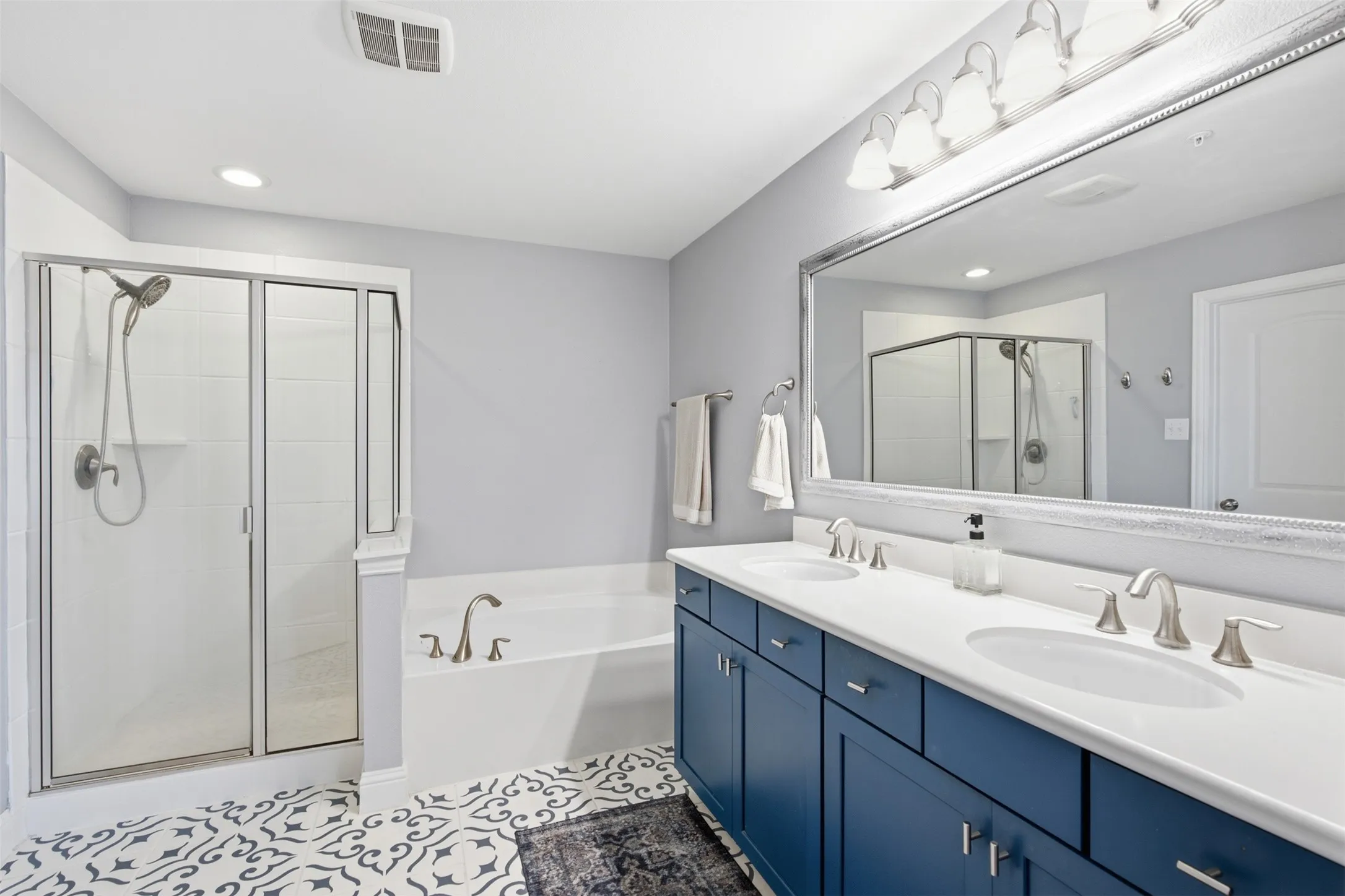Dual vanity with undermount sinks and blue cabinetry, large framed mirror, walk-in shower with glass enclosure, built-in soaking tub, and decorative floor tile