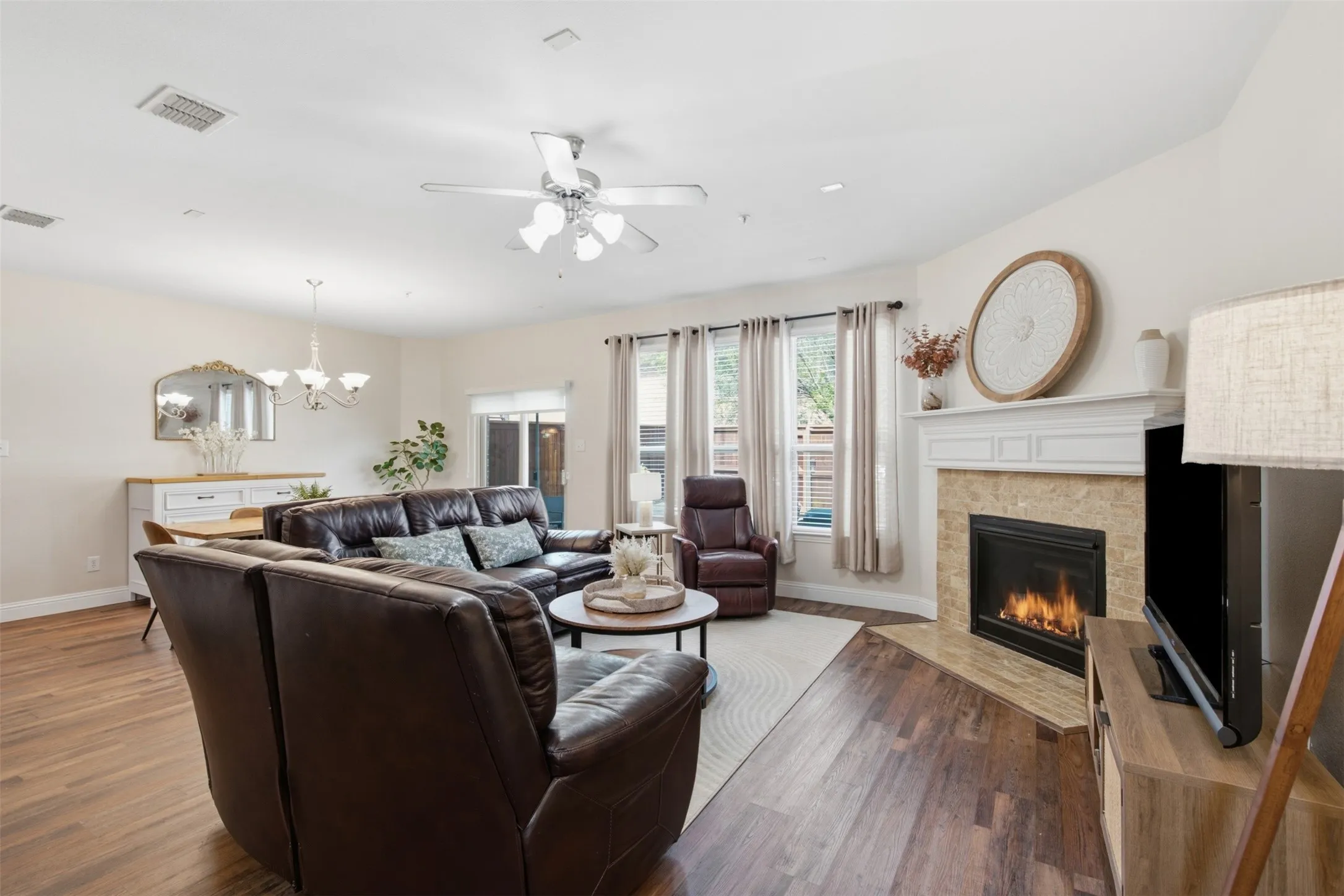 Corner fireplace with a stone surround and mantel, wood-finish flooring, ceiling fan, and multiple windows with curtains