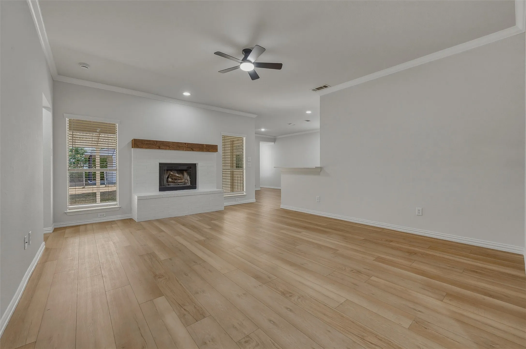 Spacious living area featuring wood-finish flooring, a white-painted brick fireplace with a wood mantle, and a ceiling fan