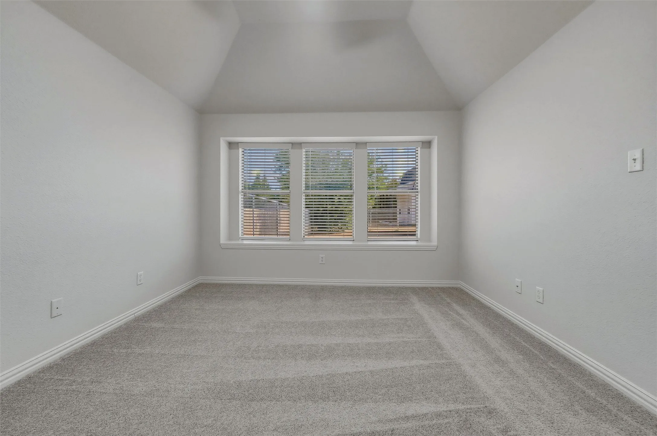 Vaulted ceiling, triple window with blinds, light-toned carpeting, and neutral wall tones