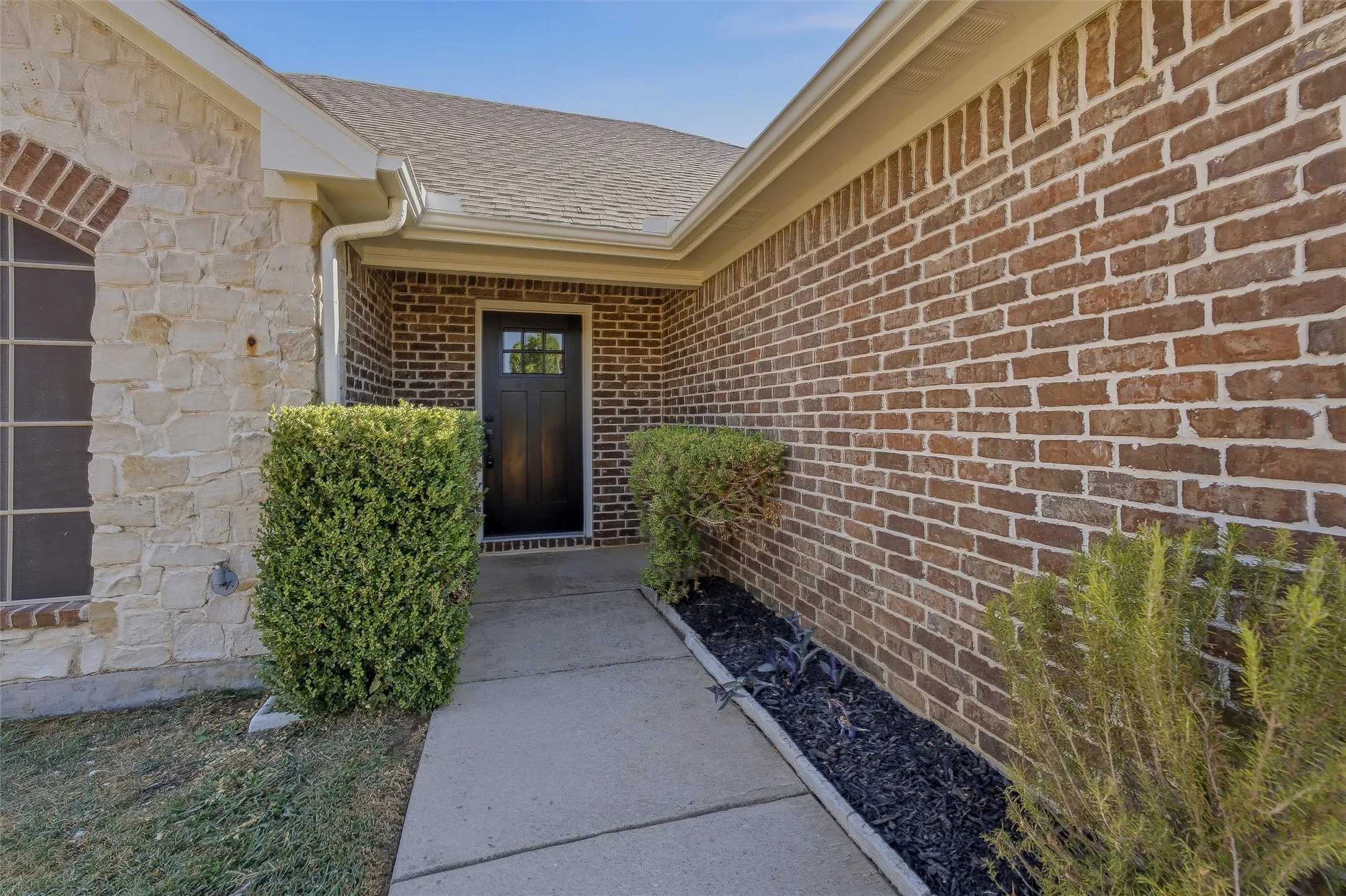 Brick and stone veneer exterior with a dark entry door, concrete walkway, and landscaped planting beds