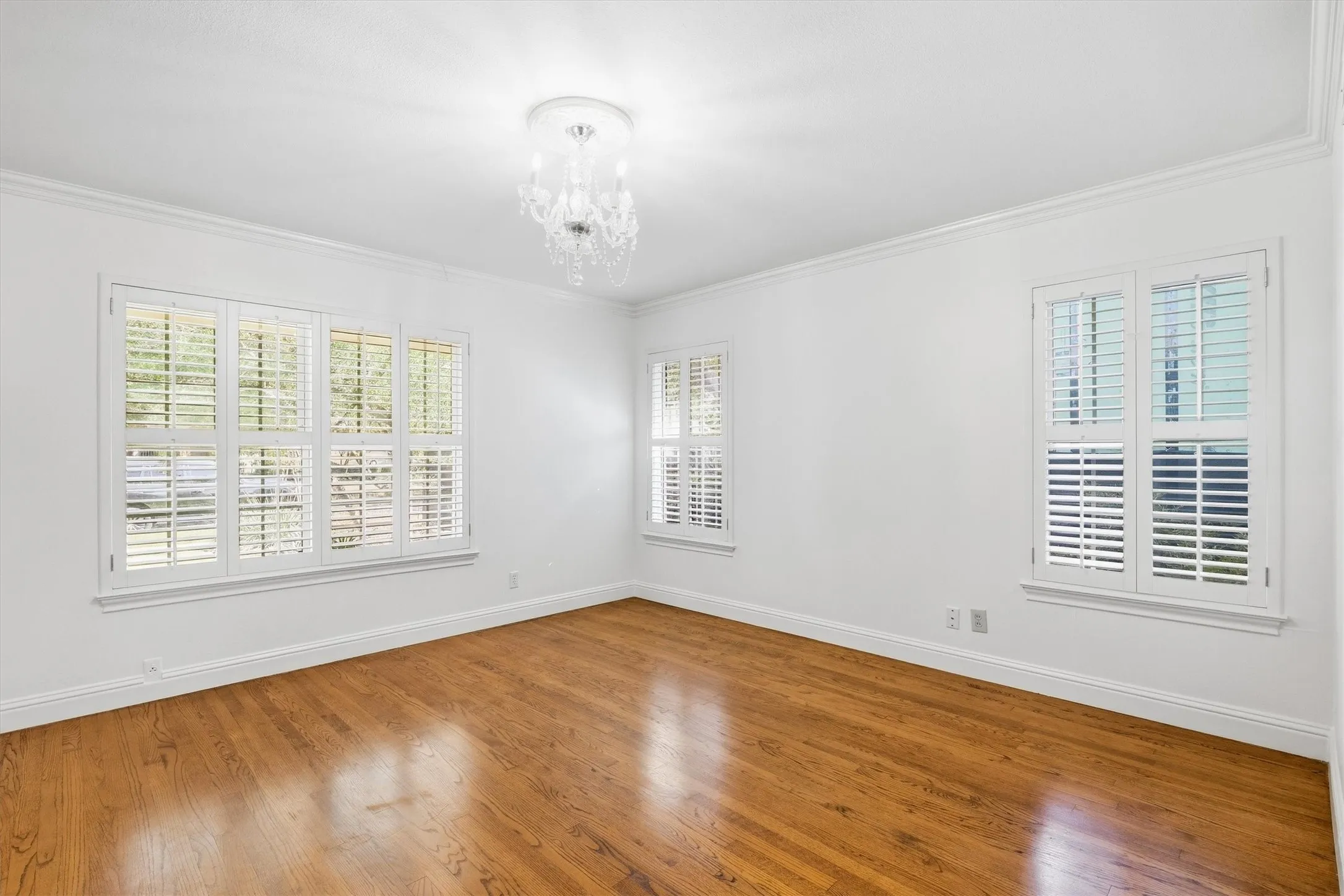 Wood-finish flooring, multiple windows with white plantation shutters, crown molding, and a decorative ceiling light fixture