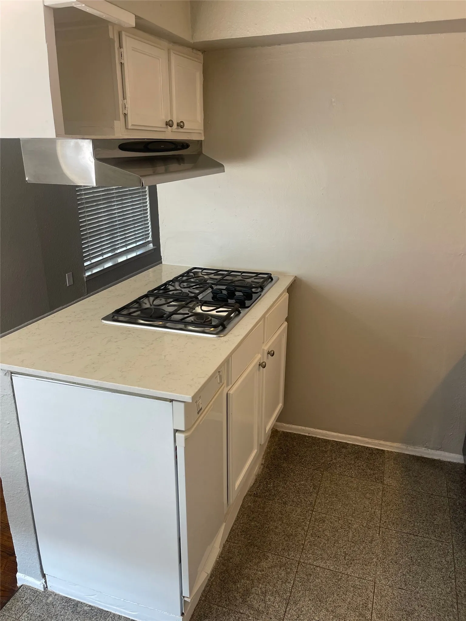 White cabinetry with gold-tone hardware, stainless steel range hood, built-in gas cooktop, light-toned countertop, and tiled flooring