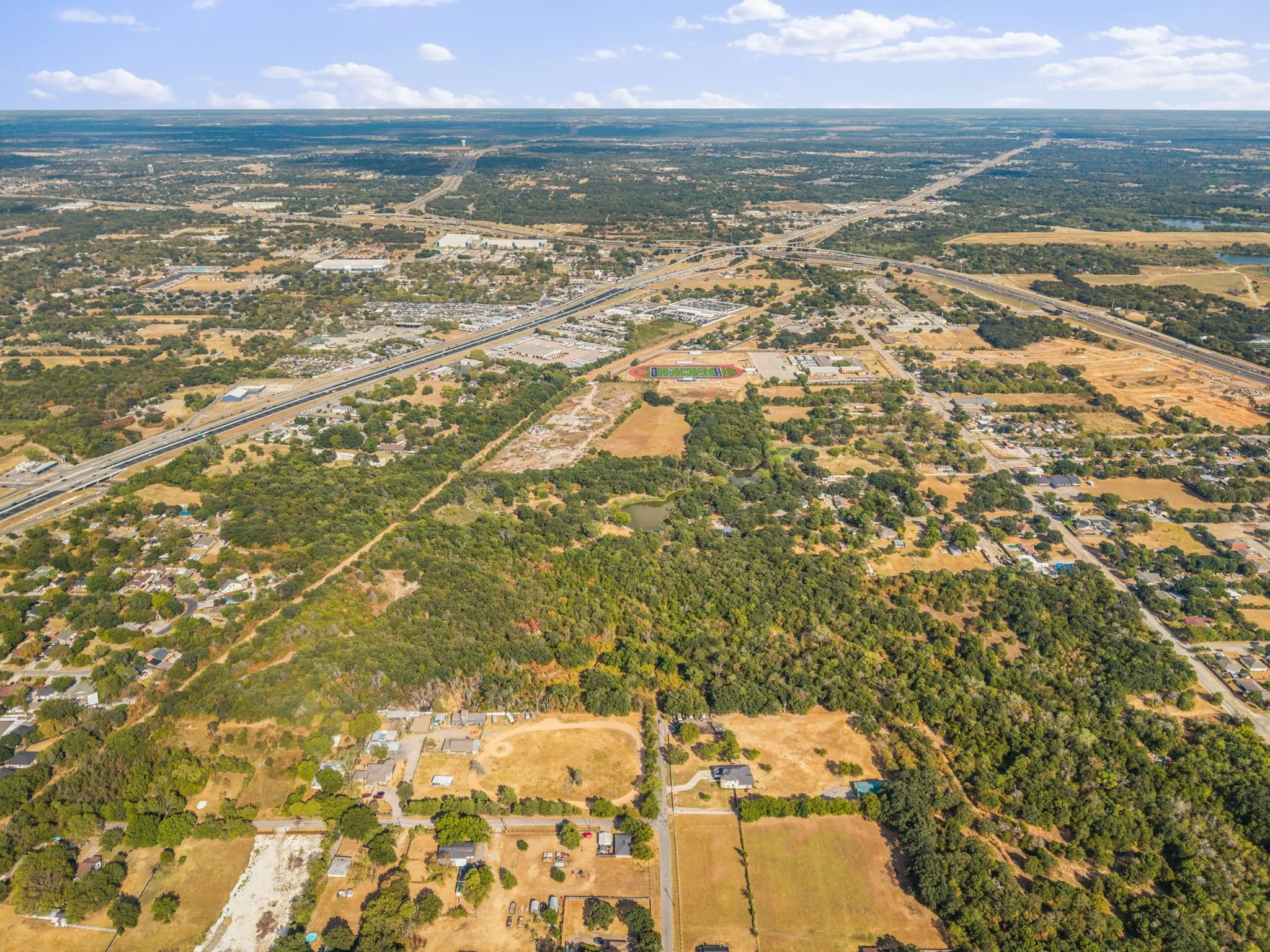 Expansive aerial perspective featuring a diverse landscape with residential areas, wooded parcels, and transportation infrastructure