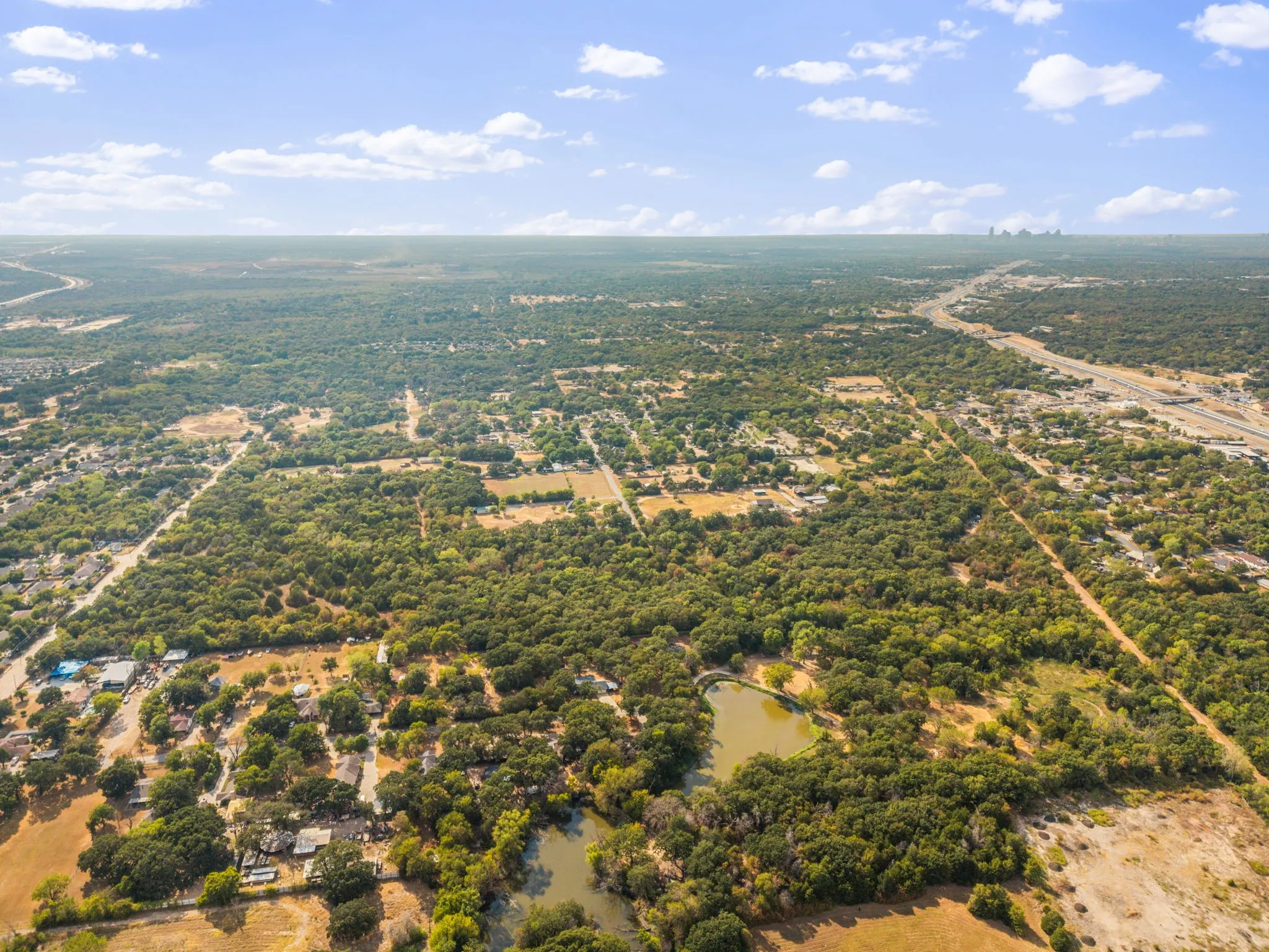 Expansive aerial showcasing a verdant landscape with mature trees, a natural pond, and distant roadways