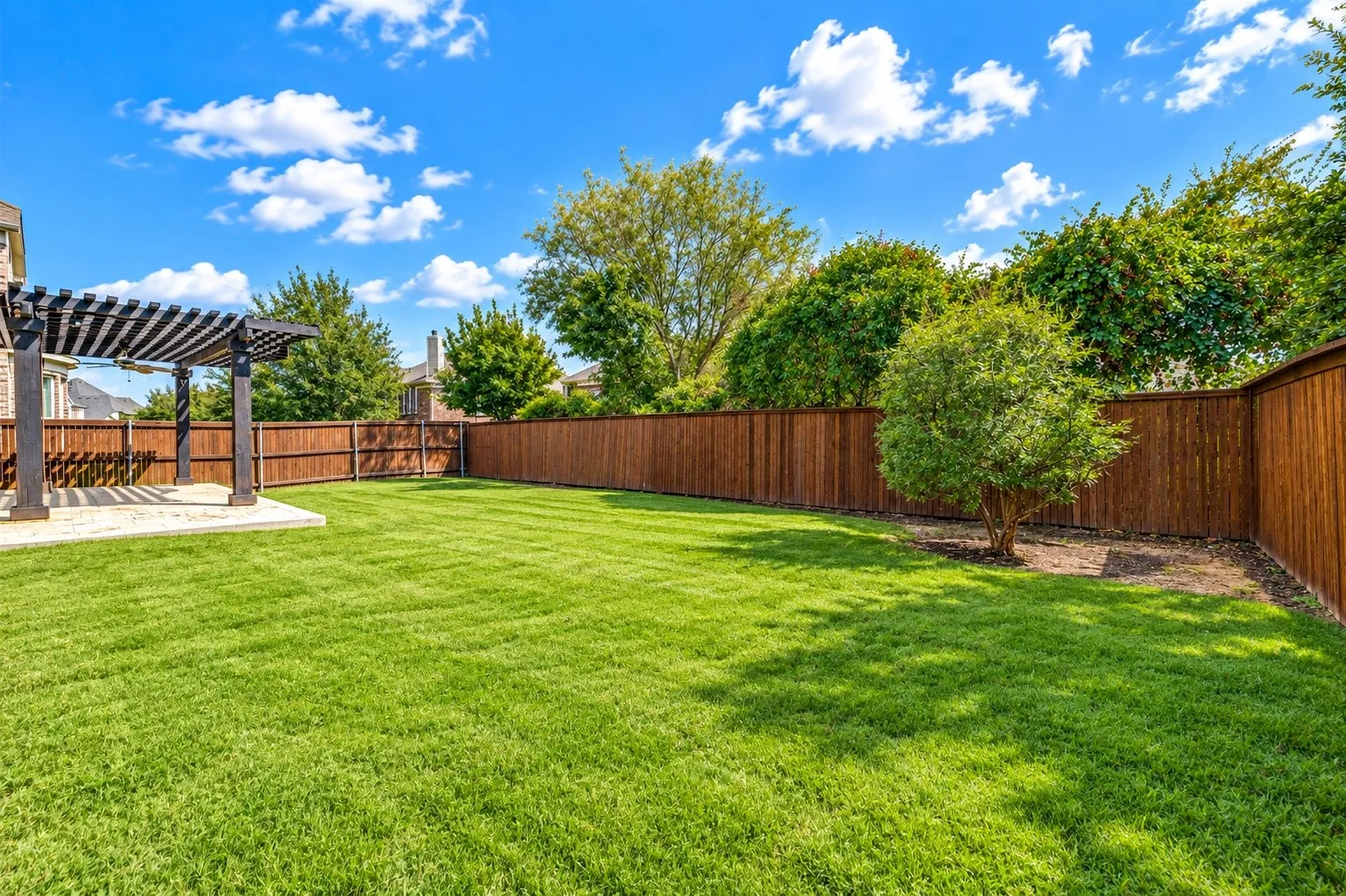 Expansive green lawn with a perimeter wood fence, mature landscaping, a concrete patio, and a wood pergola