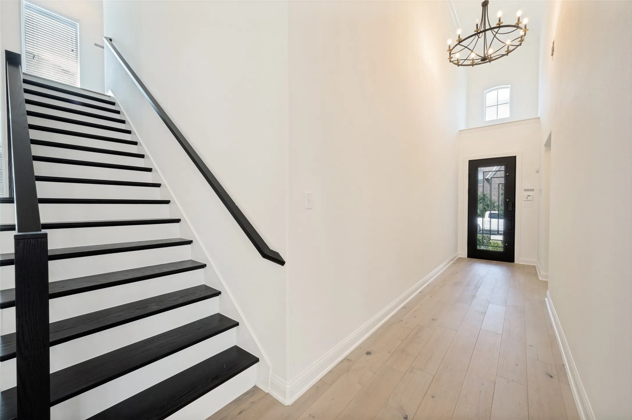 Wood-finish flooring, staircase with black treads and handrail, double-height ceiling, decorative chandelier, and an arched transom window