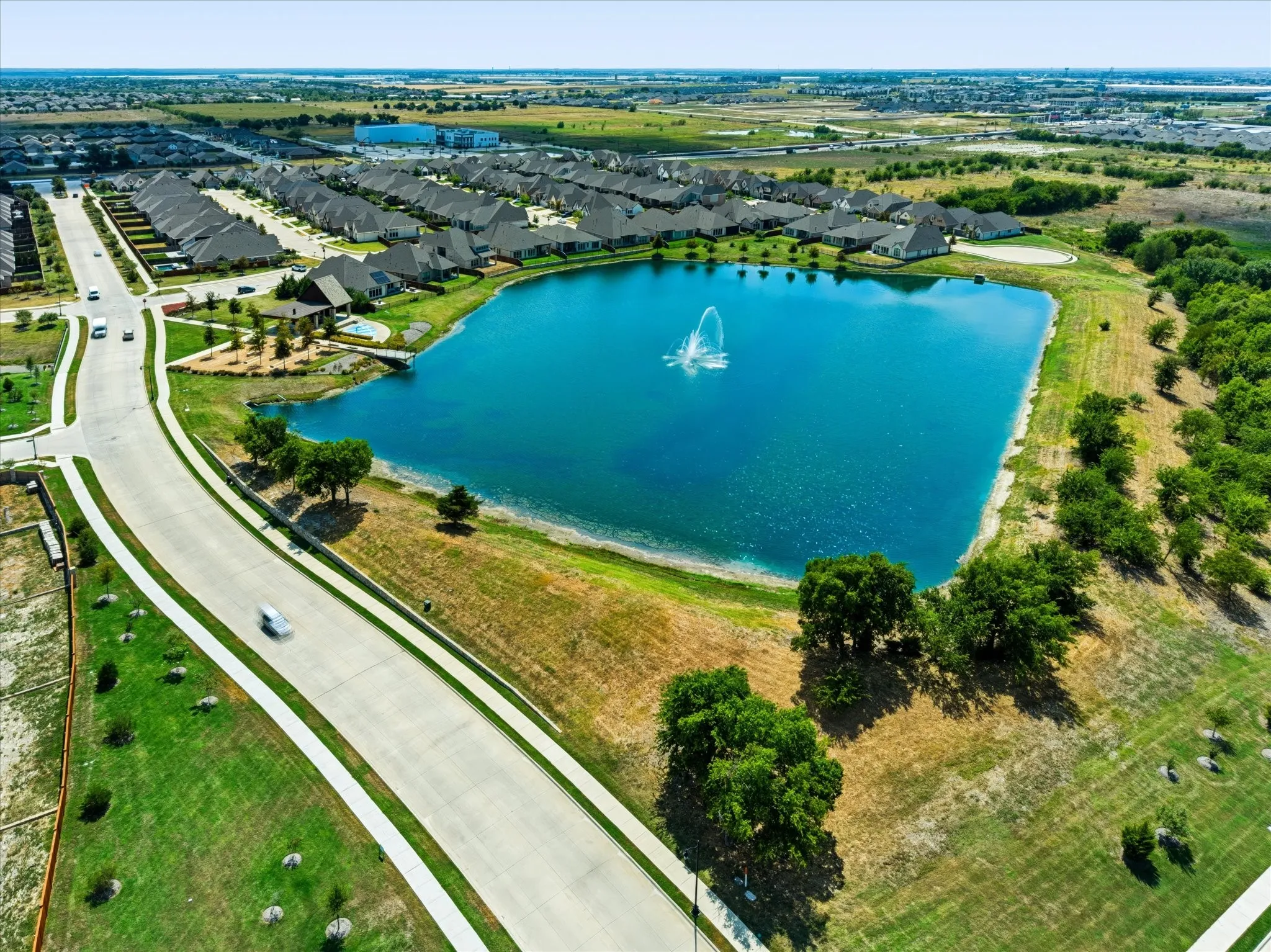 Community featuring a prominent central pond with a fountain, surrounded by residential properties, a paved road, and landscaped green spaces