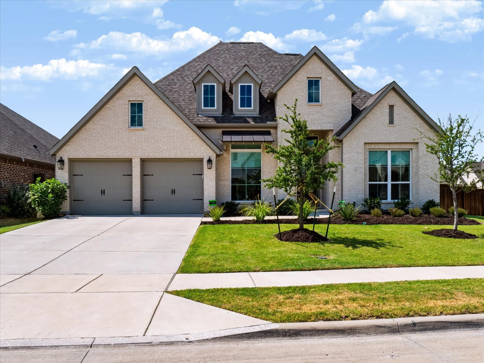 Exterior featuring a light brick facade, dark shingle roof, two-car garage, manicured lawn, and mature landscaping