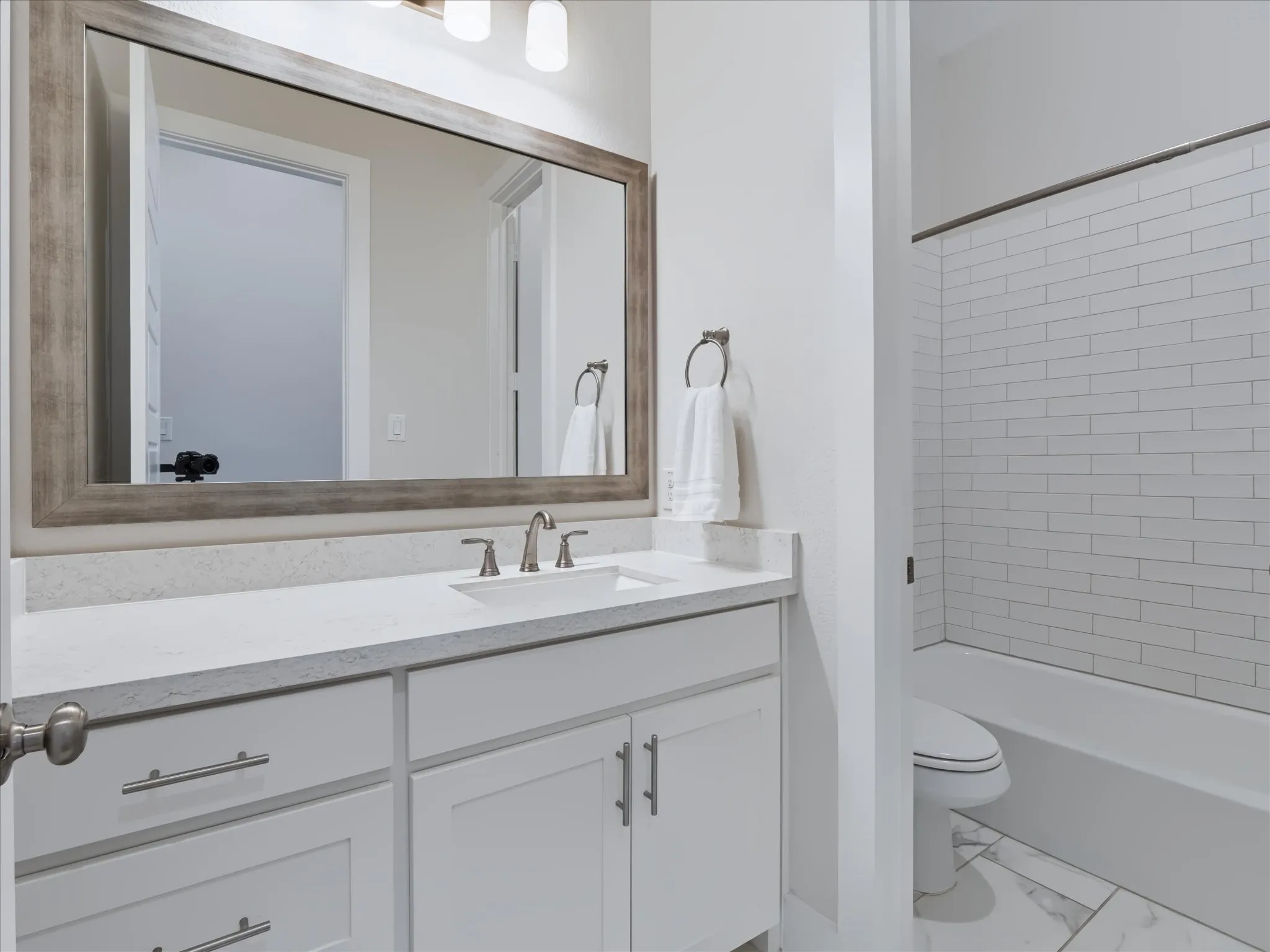 White vanity with integrated sink, rectangular wood-frame mirror, light-toned countertop, subway tile bath surround, and bath/shower combination